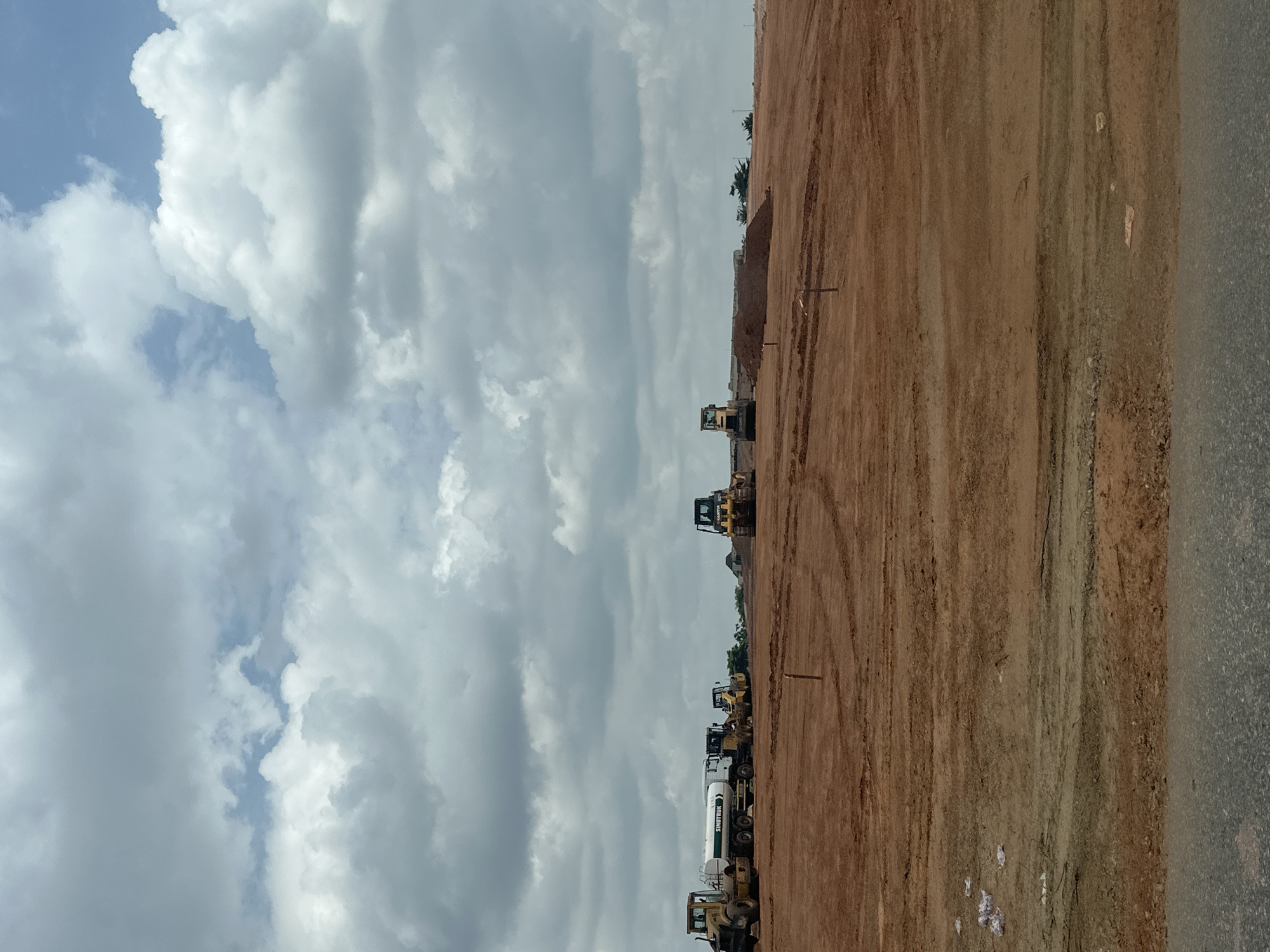 The image shows a construction site in Crescent, Nigeria, on a cloudy day. The scene is dominated by a large expanse of reddish-brown soil, with tire tracks indicating recent activity. On the left side of the frame, a line of heavy machinery is parked, including several yellow bulldozers or front-loaders and a white water truck. The truck has text on its side that reads "SWIFT TRUCK". In the background, a small pile of earth has been moved, and some sparse vegetation and buildings can be seen in the distance. The sky is filled with dramatic, white, and grey clouds, suggesting a potentially rainy or overcast day. The lighting is diffused, typical of an overcast midday. The overall impression is one of ongoing construction work, with a variety of vehicles ready for operation.