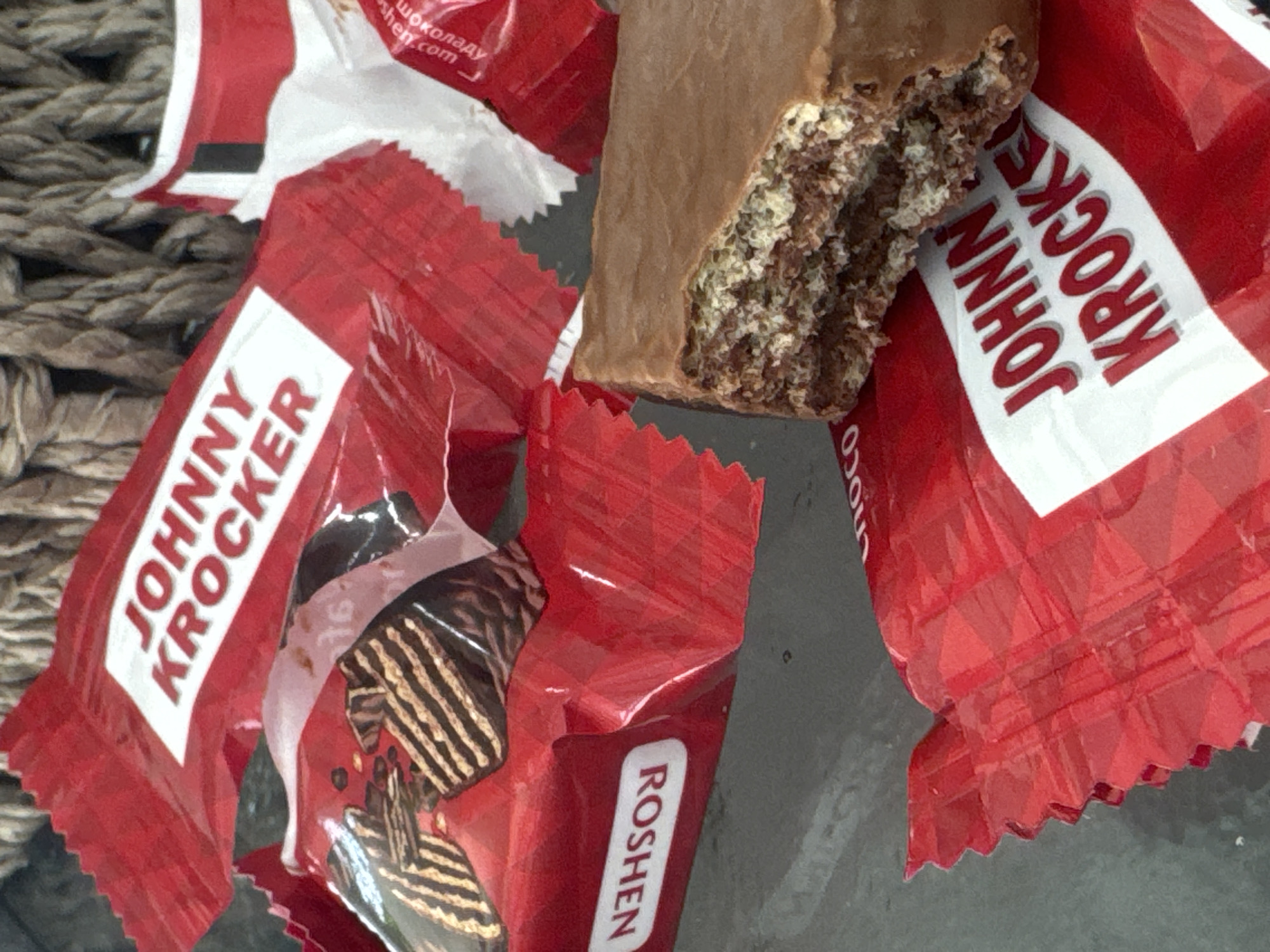 On a reflective surface, possibly glass or dark plastic, sits a partially consumed chocolate wafer bar among wrappers labeled "JOHNNY KROCKER" and "ROSHEN." The wrappers are predominantly red and white. A section of wicker is visible in the background. The scene is devoid of human presence. There is no indication that this image was taken in the city of Ramat Gan, Israel.