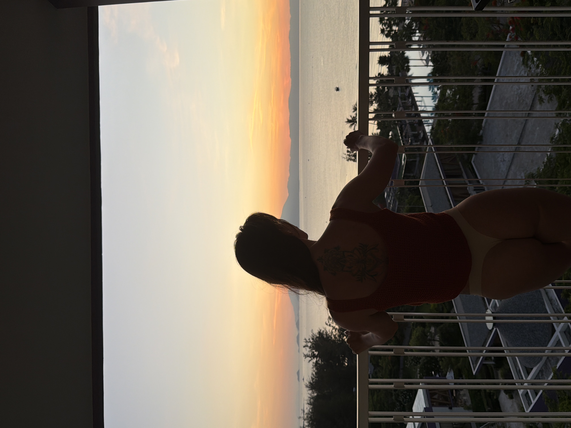 A woman stands on a balcony overlooking a body of water at sunset. She is seen from behind, wearing a red tank top and light-colored underwear. Her back features a tattoo. The sky is a gradient of orange and yellow, with distant islands or mountains visible on the horizon. Below the balcony, buildings with dark roofs and a swimming pool can be seen amidst lush greenery. The scene suggests a tropical resort or coastal area.