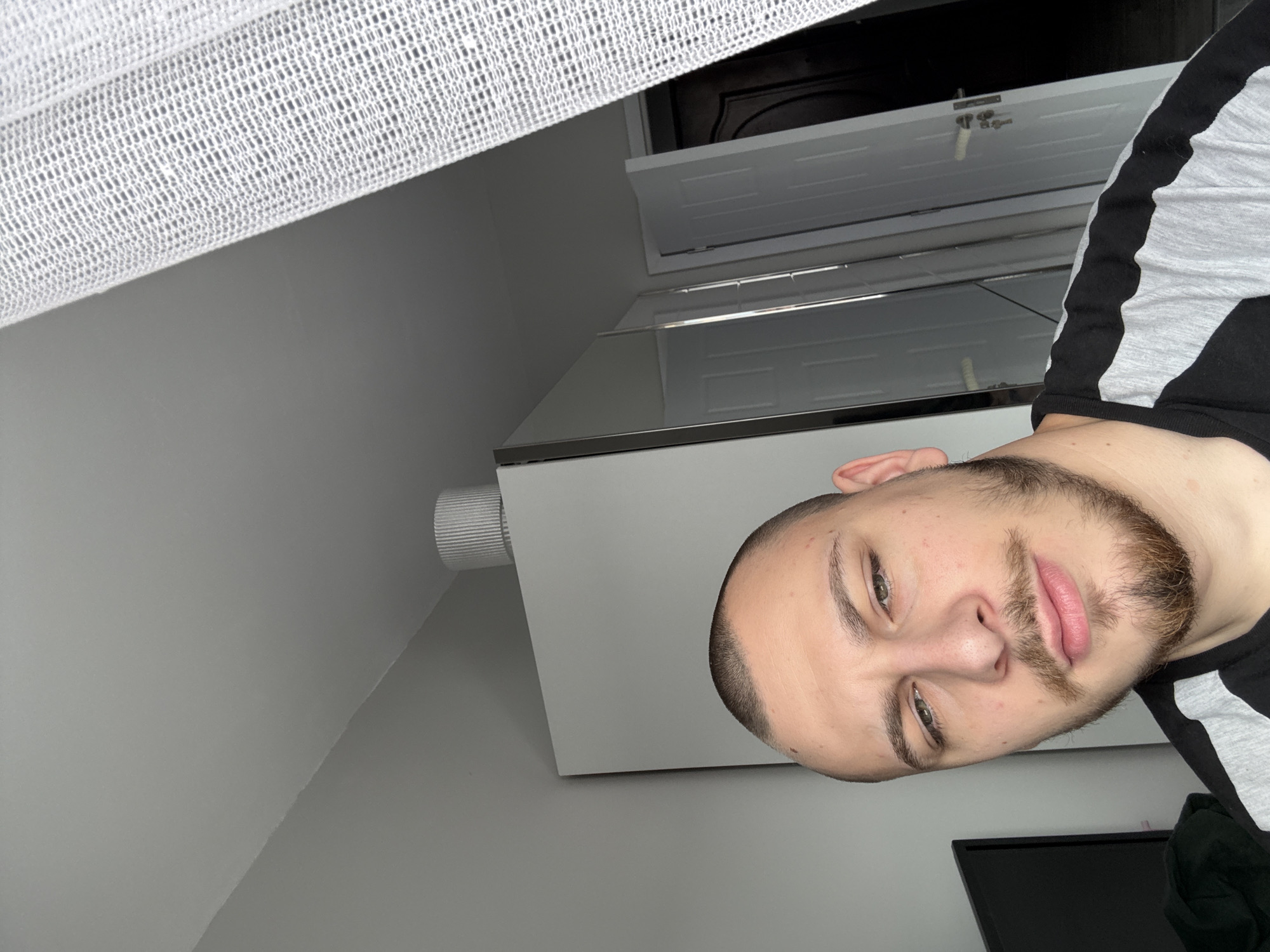 The image shows a man with short brown hair and a beard, wearing a black and gray shirt. The man is facing the camera. Behind the man is a white wall and a white cabinet. Above the cabinet are two white doors, the upper one open. Above that, an unidentifiable dark interior can be seen. A white curtain is in the top left corner of the image. There is no verifiable information to suggest that this image was taken in Stara Zagora, Bulgaria.