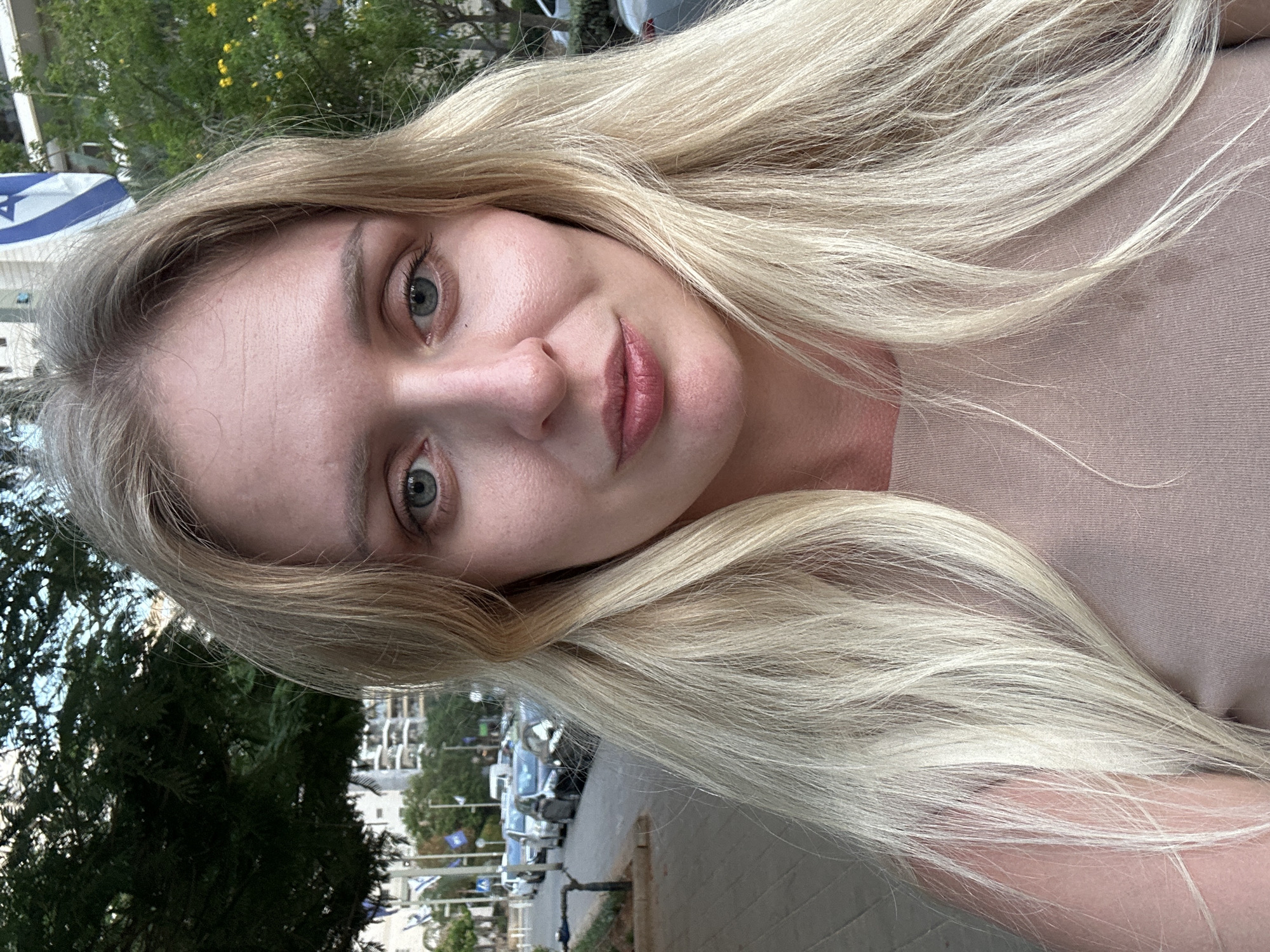 A close-up, selfie-style photograph features a young, fair-skinned woman with long, wavy blonde hair. She is looking directly at the camera, her facial features prominent. Her eyes are a light blue-green, and she has visible eyelashes and eyebrows. Her lips are slightly parted and appear to be wearing a subtle lip color. She is wearing a neutral-colored, ribbed top that exposes her shoulders and décolletage.
The background suggests an urban setting in Petah Tikva, Israel. To the left, a large, leafy green tree is visible, with a sidewalk and street lined with parked cars. Buildings with balconies are discernible in the distance. A flag, likely the flag of Israel, is visible to the right of the woman's head, consisting of a blue Star of David on a white background with two blue horizontal stripes. The overall lighting suggests it is daytime, possibly late afternoon or early evening, with natural light illuminating the scene. The image is sharp and focused on the woman's face and upper body.
