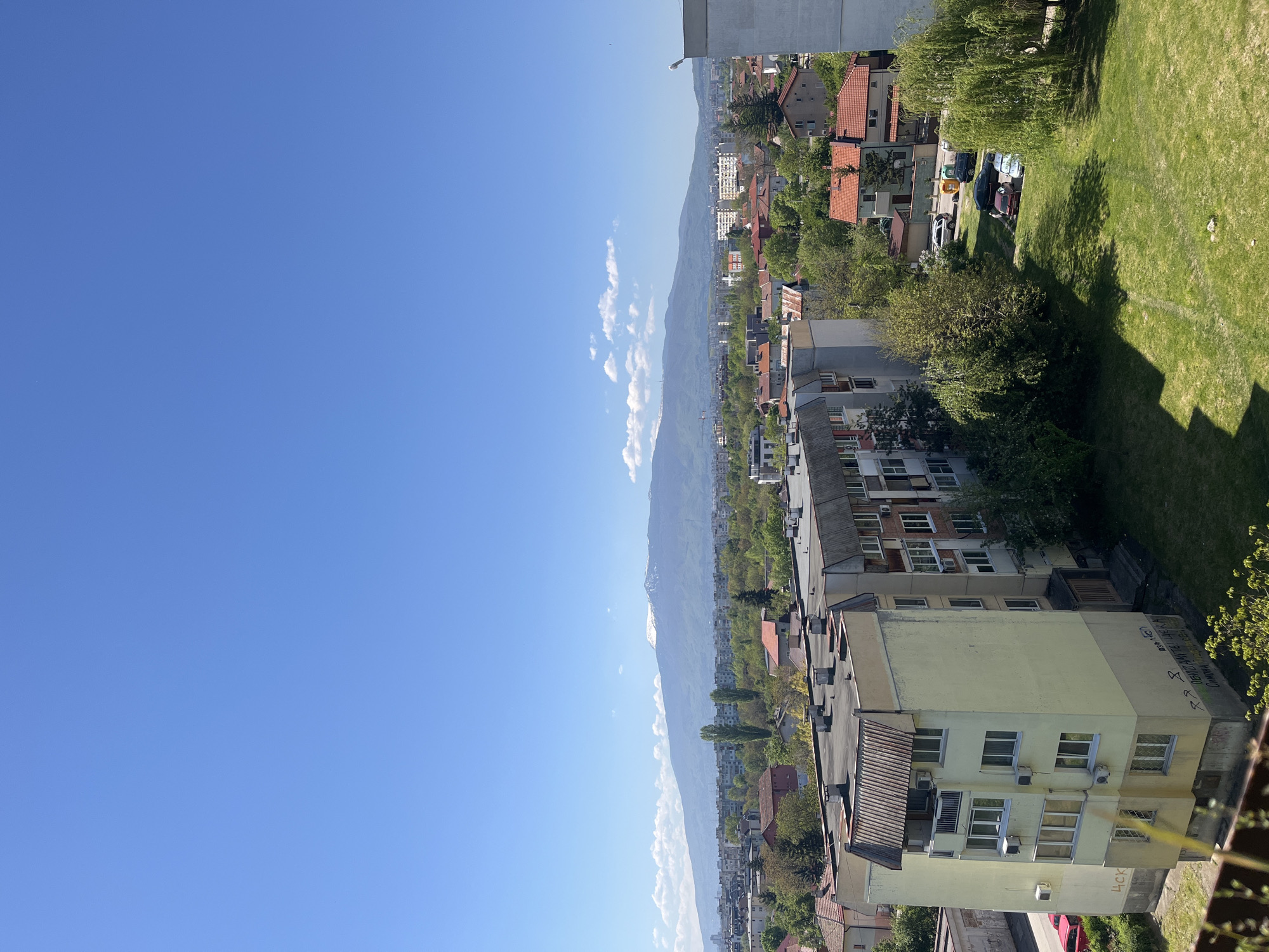 The image presents a high-angle view of an urban landscape, likely taken from a tall building in Sofia, Bulgaria. The right side of the frame features a neighborhood with residential buildings, trees, and a green lawn. In the midground, a mountain range stretches across the horizon, partially obscured by the haze. The sky is clear blue with a few scattered clouds on the left side. The overall impression is that the image captures an urban environment with scenic mountain backdrop, characteristic of Sofia.