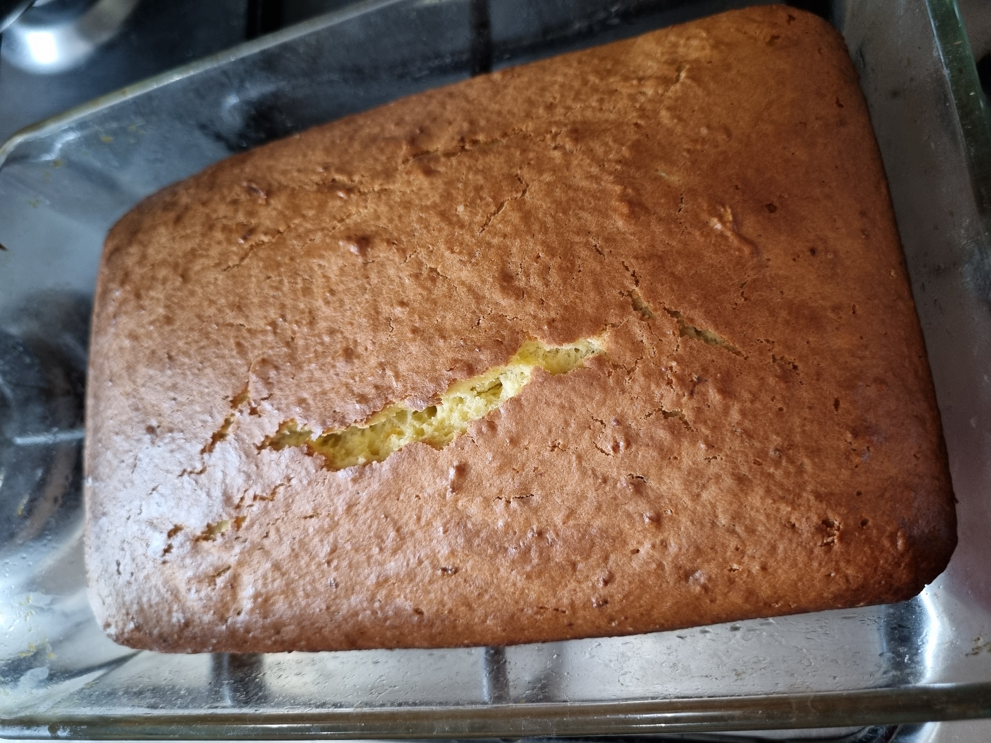 A rectangular, golden-brown baked good, likely a cake or quick bread, is centrally featured in a clear glass baking dish. The top surface of the cake has a prominent crack, revealing a lighter, yellowish interior. The crust appears textured and evenly browned. The glass dish is simple and rectangular, allowing a view of the cake's sides. It rests on what appears to be a metal grate or stovetop surface, with a dark, indistinct background suggesting an indoor kitchen environment. The scene implies the item has recently been baked and is possibly cooling. There are no people visible, and no text can be discerned in the image. The time of day is not evident from the lighting. While the image provides no specific geographical cues, the context for this media file is Chaikovsky, Russia.