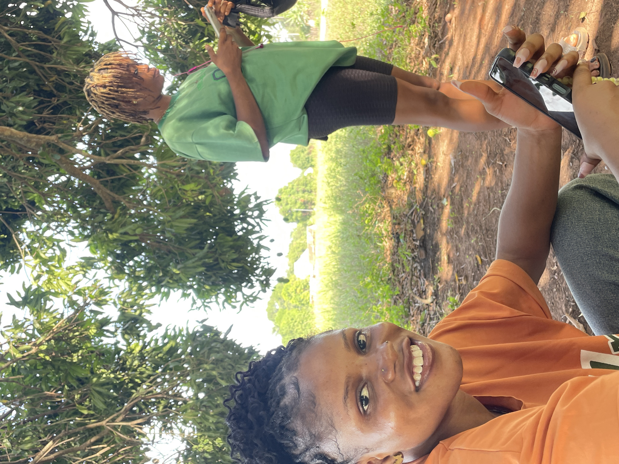 The image appears to be a selfie taken outdoors in Ogbe, Nigeria, possibly in a park or garden with trees. One woman with short, curly dark hair is in the foreground, smiling and holding a phone, presumably taking the photo. She is wearing an orange t-shirt and has piercings on her nose and ears. Another woman with blonde dreadlocks is in the background, wearing a green shirt and black shorts, also holding a phone. They seem to be taking pictures or videos of each other. The environment includes trees, grass, and a dirt path.