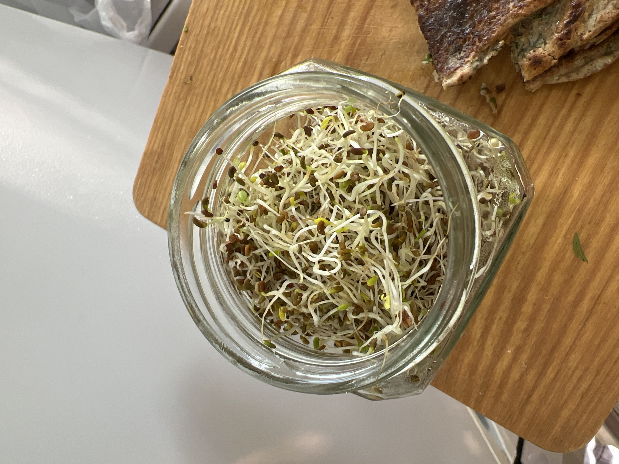 An overhead shot details an indoor kitchen or dining surface. A clear glass jar, approximately two-thirds full of mixed sprouts, rests on a light brown wooden cutting board. The sprouts display thin, white root structures with small green and yellowish cotyledons emerging from dark brown seed husks. To the right of the jar, two irregularly cut pieces of a toasted or pan-fried food item, characterized by browned exteriors and visible green specks within their lighter interior, are partially visible on the cutting board. The wooden board exhibits a discernible grain and possesses rounded edges. The background consists predominantly of a plain, light-colored wall or surface, with a blurry white object, possibly a crumpled plastic bag, visible in the upper right. A metallic edge is partially observable in the lower-left quadrant of the frame. No individuals are present.