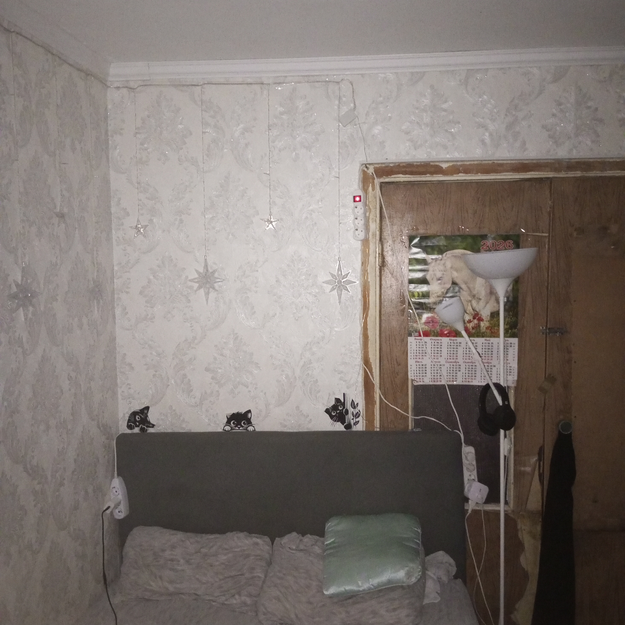 This is a medium-angle, eye-level, indoor shot of a bedroom. The room is dimly lit and appears cluttered. The walls are covered in white, floral-patterned wallpaper, and there is a white ceiling with crown molding. In the upper left corner, a plain white wall meets the floral wallpaper at a corner. Hanging from the ceiling on the floral wallpaper are several star-shaped ornaments and string lights.
In the center of the image, a grey headboard is visible, with two pillows on the bed. On top of the headboard are three black cat decals. To the right of the headboard, a floor lamp stands next to a doorway. The doorway is dark and appears to be made of wood, with some peeling paint. Inside the doorway, a calendar for the year 2026 is hanging on the wall, with an image of a donkey.
A white power strip with multiple outlets is attached to the wall to the right of the doorway, and a black cord is plugged into one of the outlets. Headphones are hanging from the floor lamp. A dark-colored garment is hanging on the wall to the right of the doorway. The overall impression of the room is one of disarray.