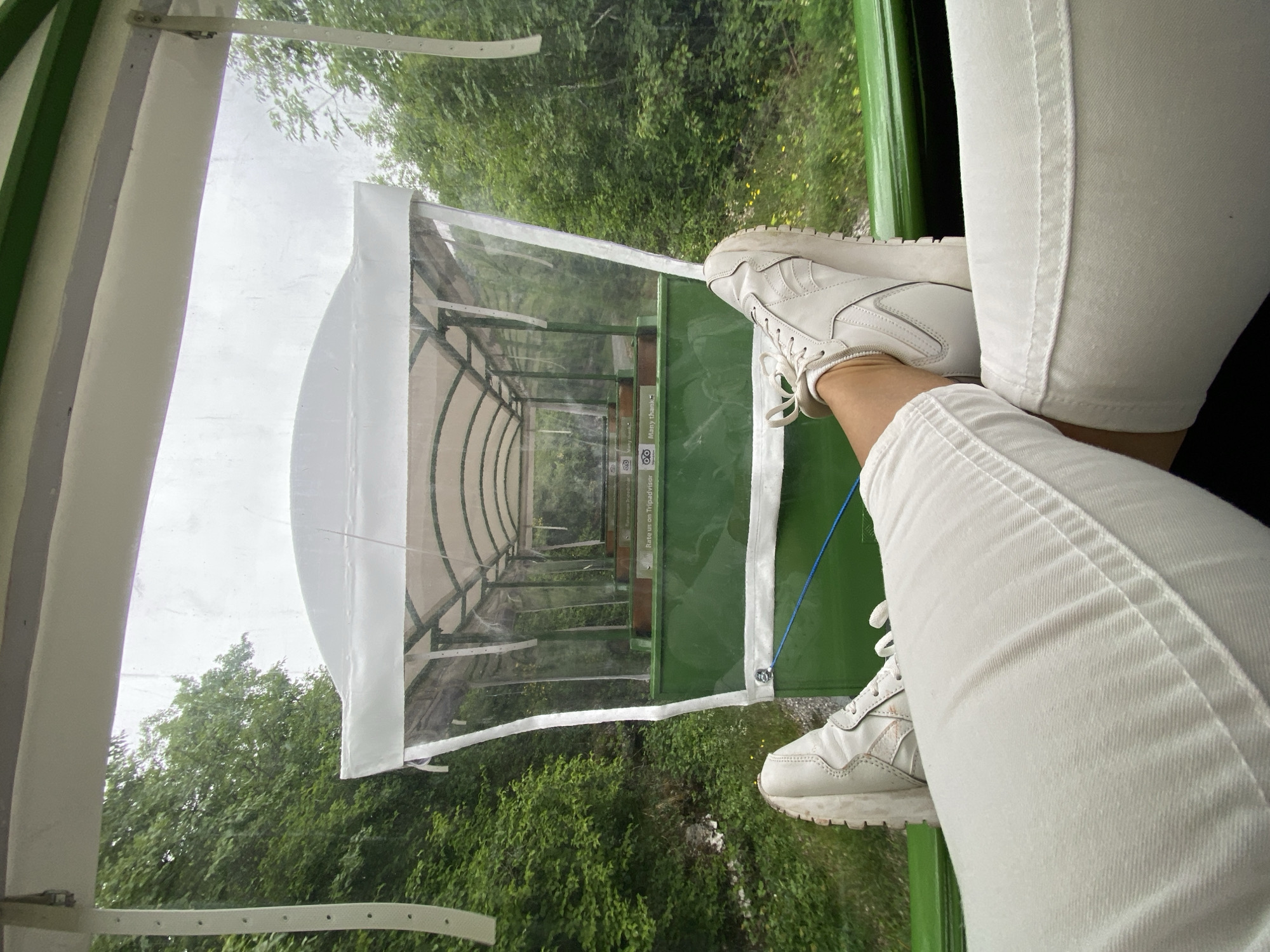 The image shows a first-person perspective from inside a small, open-air train or trolley. The viewer is wearing white pants and white sneakers and has their feet resting on the outside of the vehicle. Through the open front of the trolley, a covered walkway or structure is visible, lined with benches. Beyond the walkway, there is dense green vegetation. The presence of Tripadvisor logos suggests this is a tourist attraction, possibly in the Lipa Dobrska area of Montenegro.