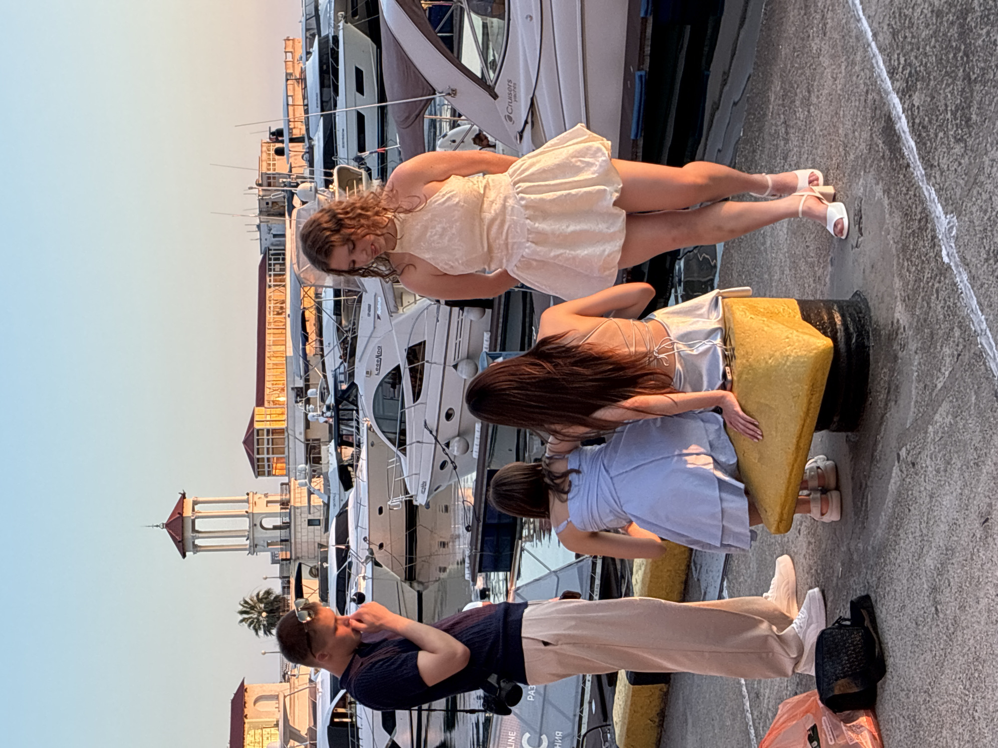 In Sochi, Russia, four young adults are gathered on a concrete pier overlooking a marina filled with white yachts. The sky is clear with a golden hue, indicating sunset.
On the left, a young man stands with his head bowed, his hand touching his face, wearing a dark short-sleeved shirt and light-colored trousers. A camera is attached to his hip.
In the center, two young women are seated on a yellow bollard. One, wearing a light blue dress, is seen from the back. The other, with long dark hair, is also facing away from the camera.
On the right, a young woman stands facing the group, wearing a short, cream-colored dress with a halter neckline. Her hair is curly and falls to her shoulders. She is wearing white heeled sandals.
In the background, a multi-story building with a red-tiled roof and white columns stands prominently, along with other buildings and numerous yachts docked in the water. The overall scene depicts a casual gathering during a warm evening.