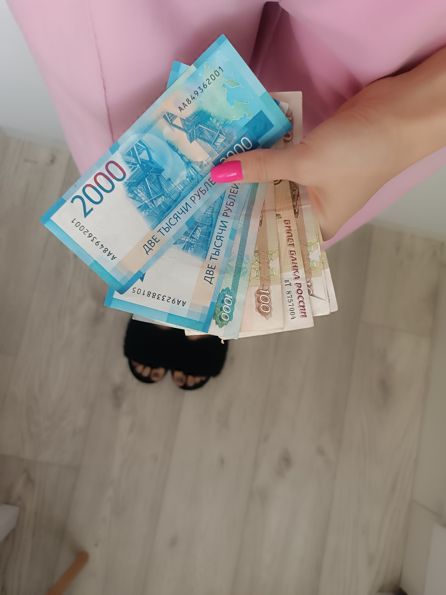 A person wearing pink pants holds Russian currency. The currency is mostly 2000 rubles, with some smaller bills. The floor and background appear to be in an indoor setting. The location is Unknown, Unknown. The person is wearing black furry slippers.