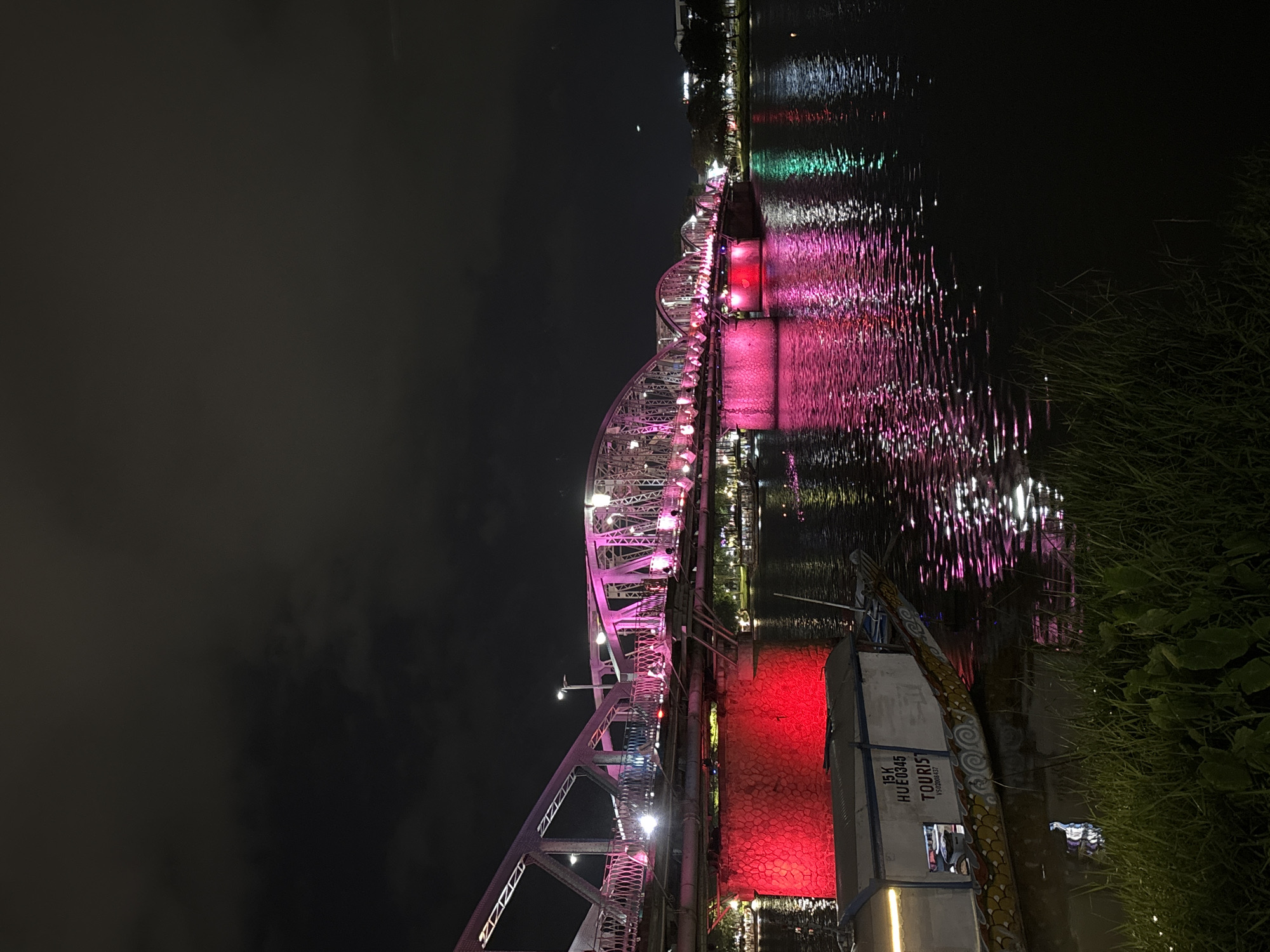 A long exposure photograph captures the Truong Tien Bridge in Phú Xuân Ward, Vietnam, at night. The bridge is illuminated with pink and red lights, casting vibrant reflections on the dark water below. The water's surface is disturbed, creating shimmering streaks of light. In the foreground, a traditional Vietnamese dragon boat is moored, with its white cabin displaying Vietnamese text including "TOURIST". Lush green grass lines the bank of the river. The sky is dark with some visible clouds. The overall scene conveys a sense of nocturnal beauty and tranquility.