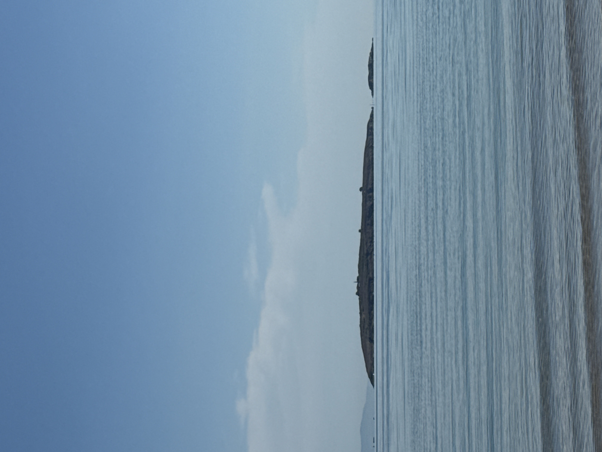 The image shows a panoramic view of the sea with islands in the distance under a clear blue sky. The water is calm with subtle ripples, reflecting the light. In the mid-ground, a long, dark island with a few structures on top, possibly a lighthouse and some buildings, is visible. To the right of the main island, a smaller rocky outcrop emerges from the water. In the far distance, the hazy silhouette of a larger landmass can be seen on the left. The foreground shows the gentle lapping of waves on a sandy shore. The scene depicts a serene coastal landscape.