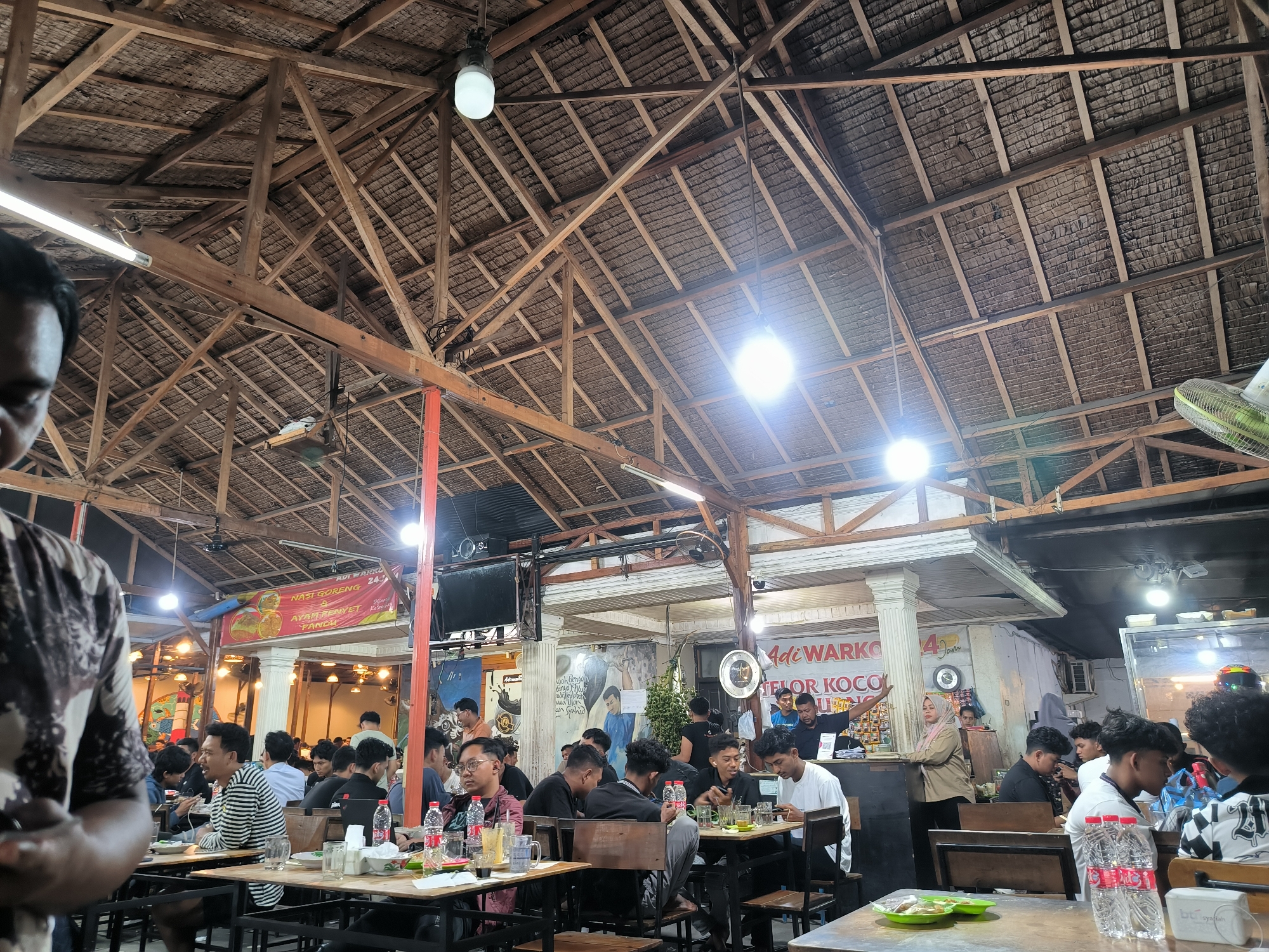 This image captures a lively night scene inside a busy, open-air eatery or warung in Banda Aceh, Indonesia. The setting features a rustic, high-peaked roof constructed from prominent wooden beams and what appears to be a thatched or woven material, characteristic of traditional Indonesian architecture. Numerous bright bare lightbulbs and fluorescent lights illuminate the space, suggesting it is nighttime.
The scene is bustling with people, predominantly men, seated at simple wooden tables and chairs. They are engaged in dining, drinking, socializing, and many are seen using their mobile phones. Some individuals are visible behind a counter, possibly staff members serving customers. The atmosphere is casual and social.
Notable objects include green plates with food, bottled water, glasses, and a ceiling fan. In the background, there's a television screen mounted, and various signs. Visible text on banners and signs, in Indonesian, indicates the name of the establishment, "ADI WARKO," with one sign specifying "24 Jam" (24 hours), suggesting it's open around the clock. Other text mentions menu items like "NASI GORENG & AYAM PENYET PANCH" (Fried Rice & Smashed Chicken) and "TELOR KOCO" (likely 'Telur Kocok,' a type of egg drink). A small text "syafah" is visible on a bottle. The overall impression is that of a popular local spot for meals and social gatherings.