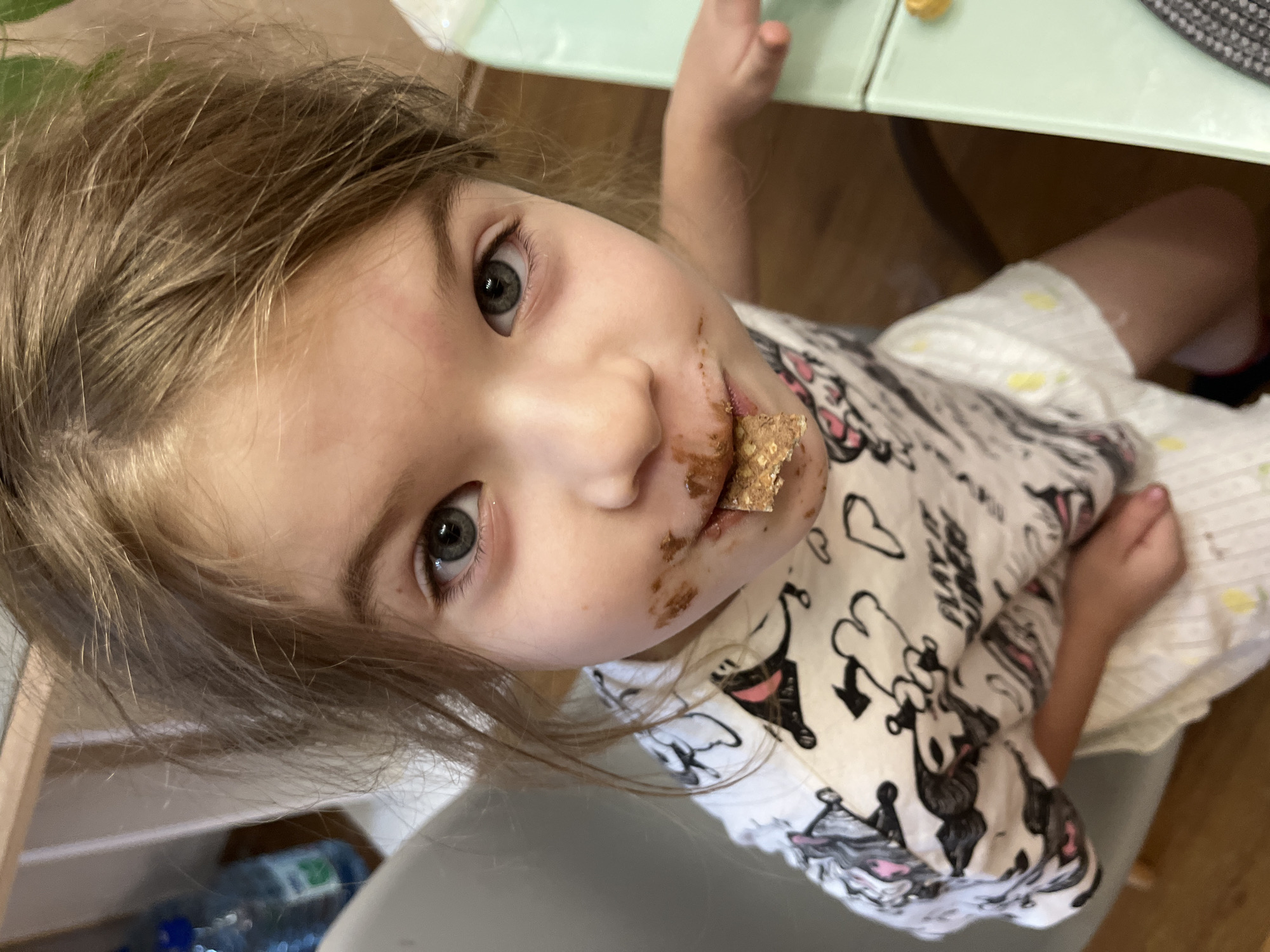 A young child with light brown hair and gray eyes is looking up at the camera. The child's face has chocolate smeared around their mouth, and they are holding a piece of food covered in chocolate. The child is wearing a white t-shirt with black and white cartoon characters printed on it. The child is seated at a table with a light green top. A water bottle is visible in the lower left corner. The location is presumed to be indoors, but the specific city of Sochi, Russia cannot be confirmed from the image alone.