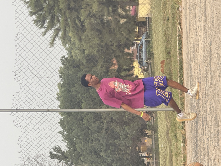 An outdoor scene in Umunekwu, Nigeria, features a young Black male standing on a paved court. He is positioned in a mid-stride or dynamic pose, leaning slightly forward and to his right, with his left foot elevated. He wears a magenta short-sleeved t-shirt with a multi-colored graphic, purple basketball shorts displaying 'LAKERS' in yellow text, and light-toned athletic shoes. A yellow wristband is on his left wrist. A basketball is partially visible on the ground in the lower-left foreground. A grey wire mesh fence, with an uneven top edge and a vertical metal post, spans the midground. Beyond the fence, a dense collection of green trees fills the background. In the further distance, a light blue or grey vehicle and faint building structures are visible under an overcast sky.
