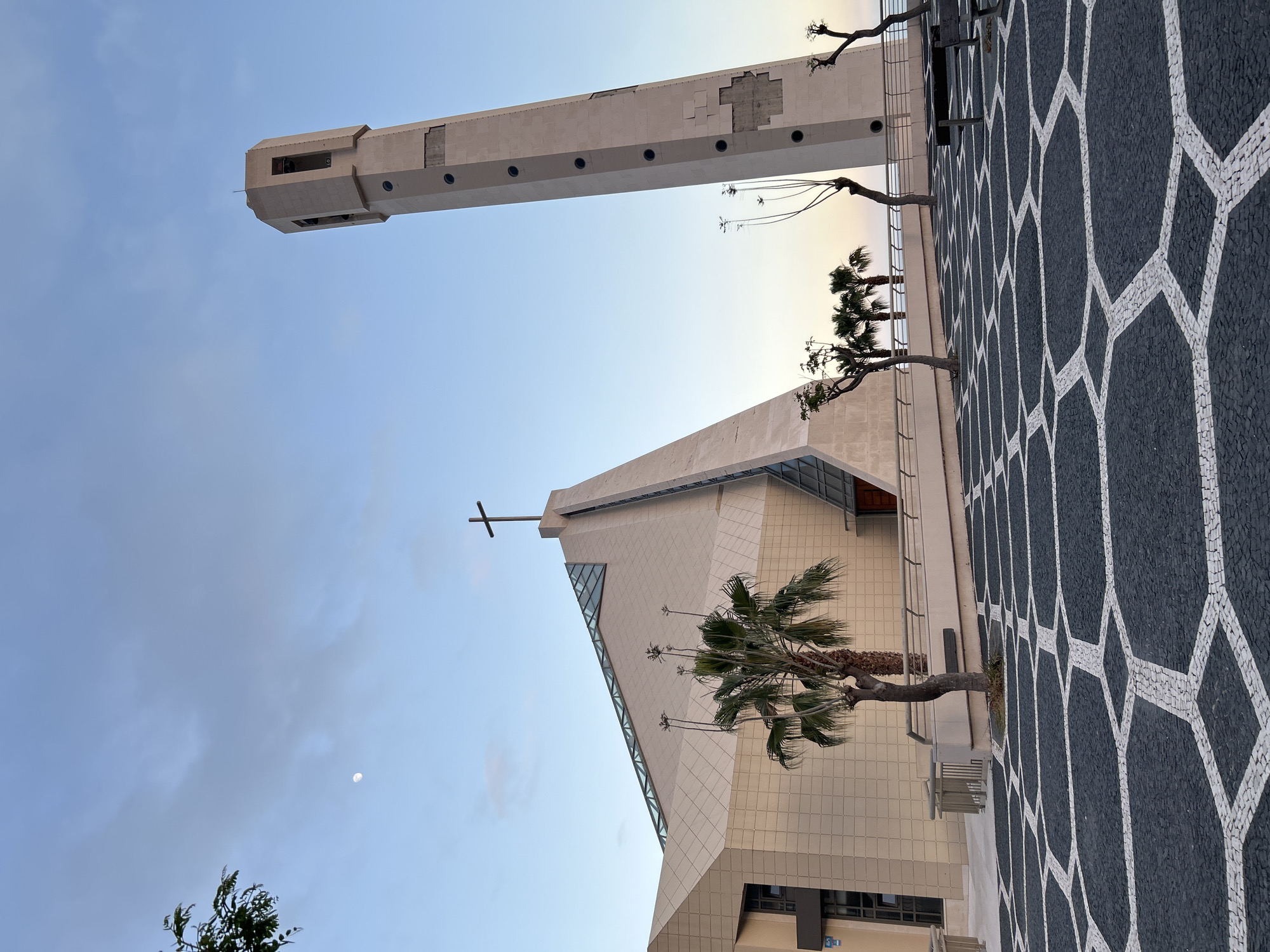 The image shows the Igreja de Santa Cecília (Church of Saint Cecilia) in Câmara de Lobos, Portugal. The church is a modern building with a tall, narrow bell tower. A cross is mounted on the facade. A geometric mosaic floor pattern is visible. Palm trees and other vegetation are included in the frame. The sky is light blue.