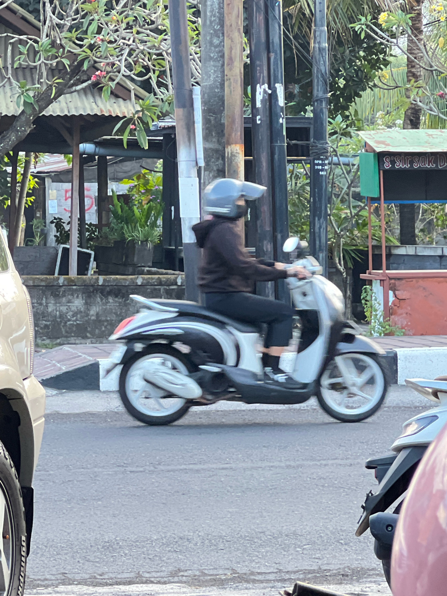 The image captures a scene on a street in Denpasar, Indonesia. A person wearing a dark hoodie and a helmet is riding a silver and black scooter. The scooter has white rims on its wheels and a silver body. The rider's legs are visible, clad in black pants and white shoes. In the foreground, a portion of a light-colored vehicle is visible on the left. Behind the scooter, there is a gray concrete wall and a row of potted plants. A sign with green and red accents is visible on the right, with text that includes "S' Sir Sak D." Electrical poles and wires are present in the background, along with trees and some structures with corrugated metal roofs. The street surface is asphalt.