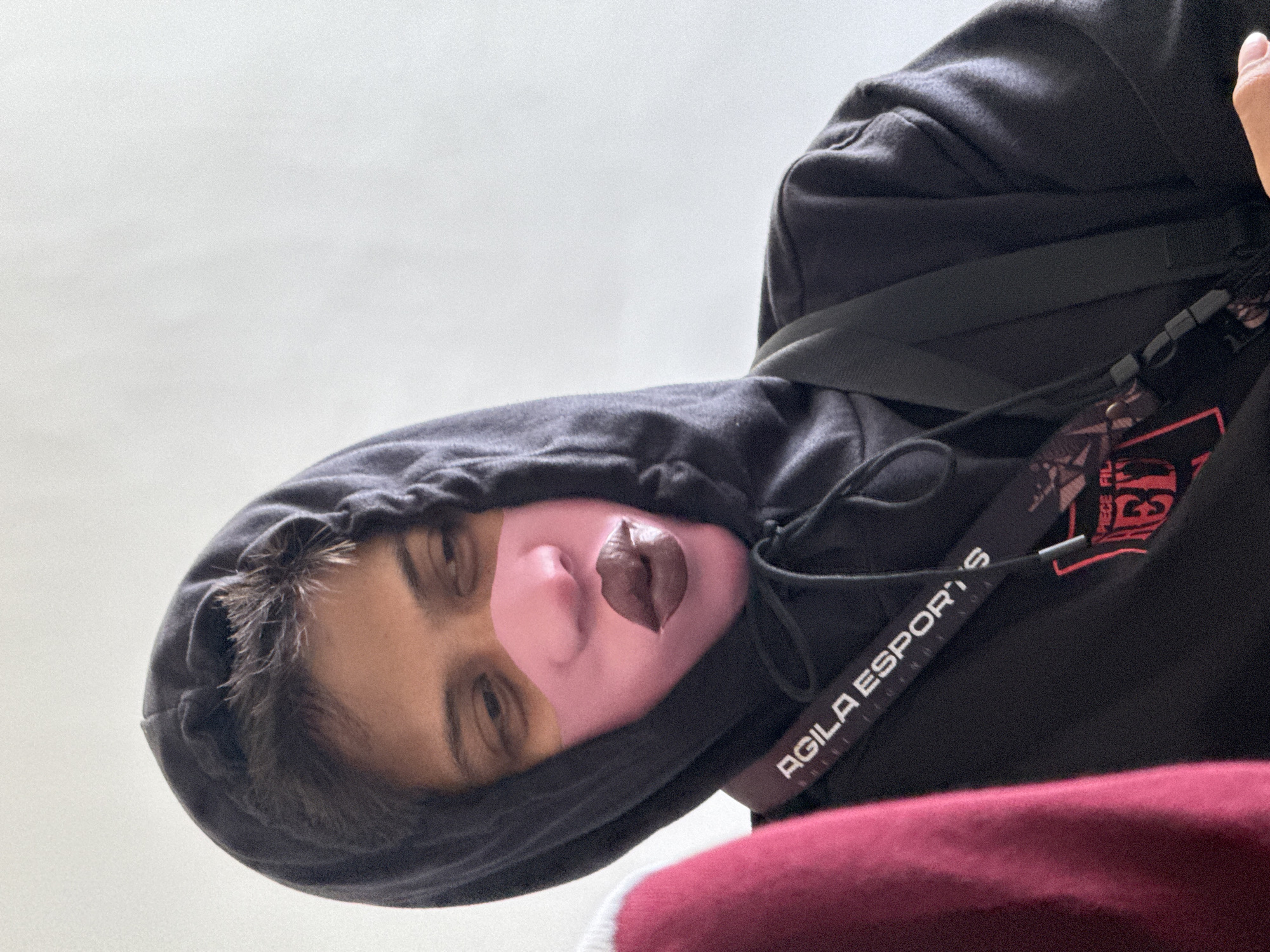 A person wearing a black hoodie with a strap and an ID with "AGILA ESPORTS" visible is visible in the image. The person's face appears to have a pink mask applied, with dark-colored lips. The background appears to be a white or light-colored surface. Without more context, I cannot determine if this image was taken in Davao City, Philippines, or anywhere else.