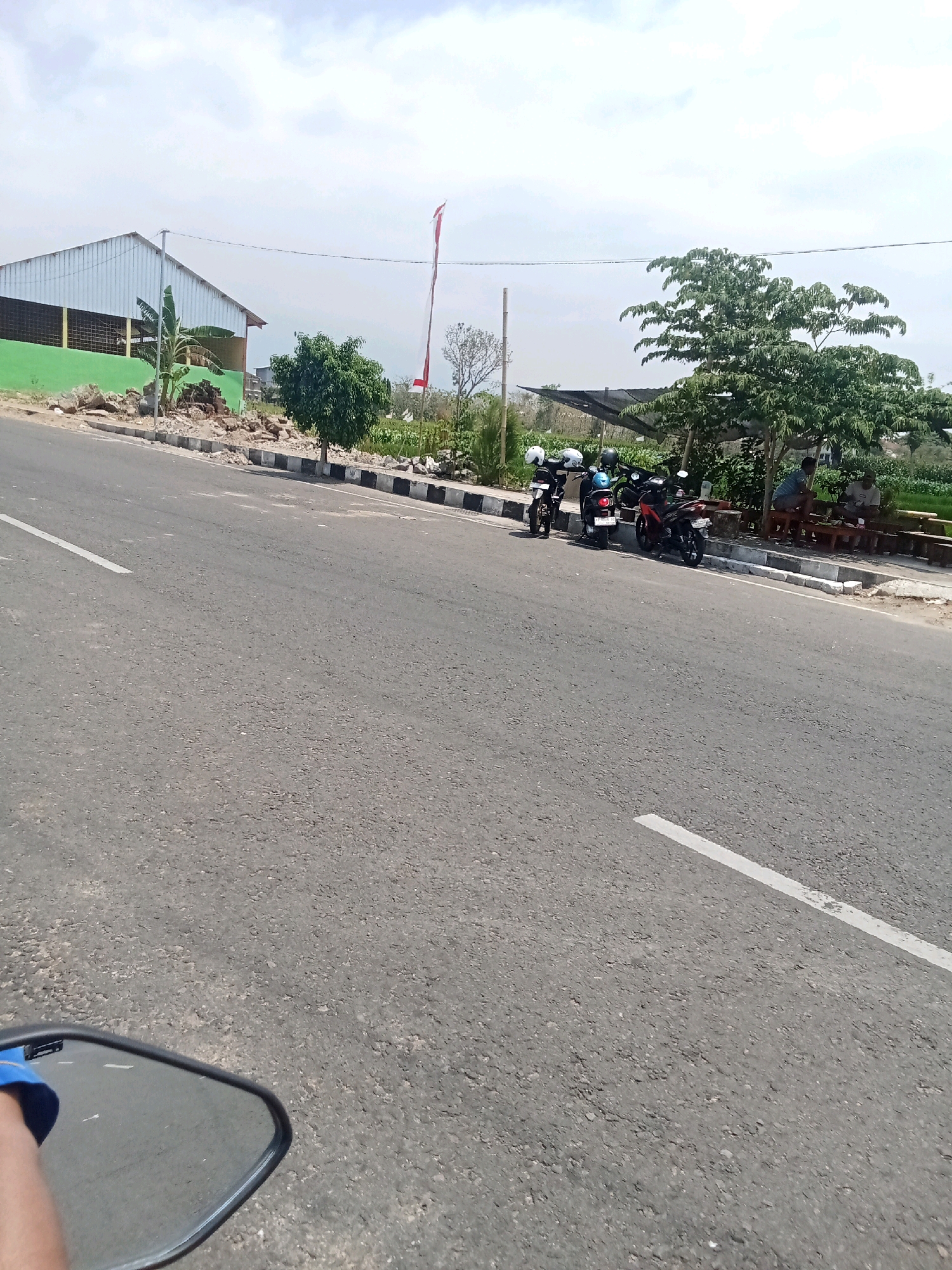 The image depicts a roadside scene in Sukowinangun, Indonesia. A paved asphalt road with a white line marking dominates the foreground. On the left side of the road, a motorcycle mirror reflects a hand wearing a blue sleeve. To the right of the road, there are several motorcycles parked. Behind the motorcycles, a group of people are seated on wooden benches under a tree, appearing to be socializing or resting. In the background, a large, light-colored building with a green base is visible, and a flag with red and white stripes flies from a pole. The surrounding environment includes trees, vegetation, and a cloudy sky.