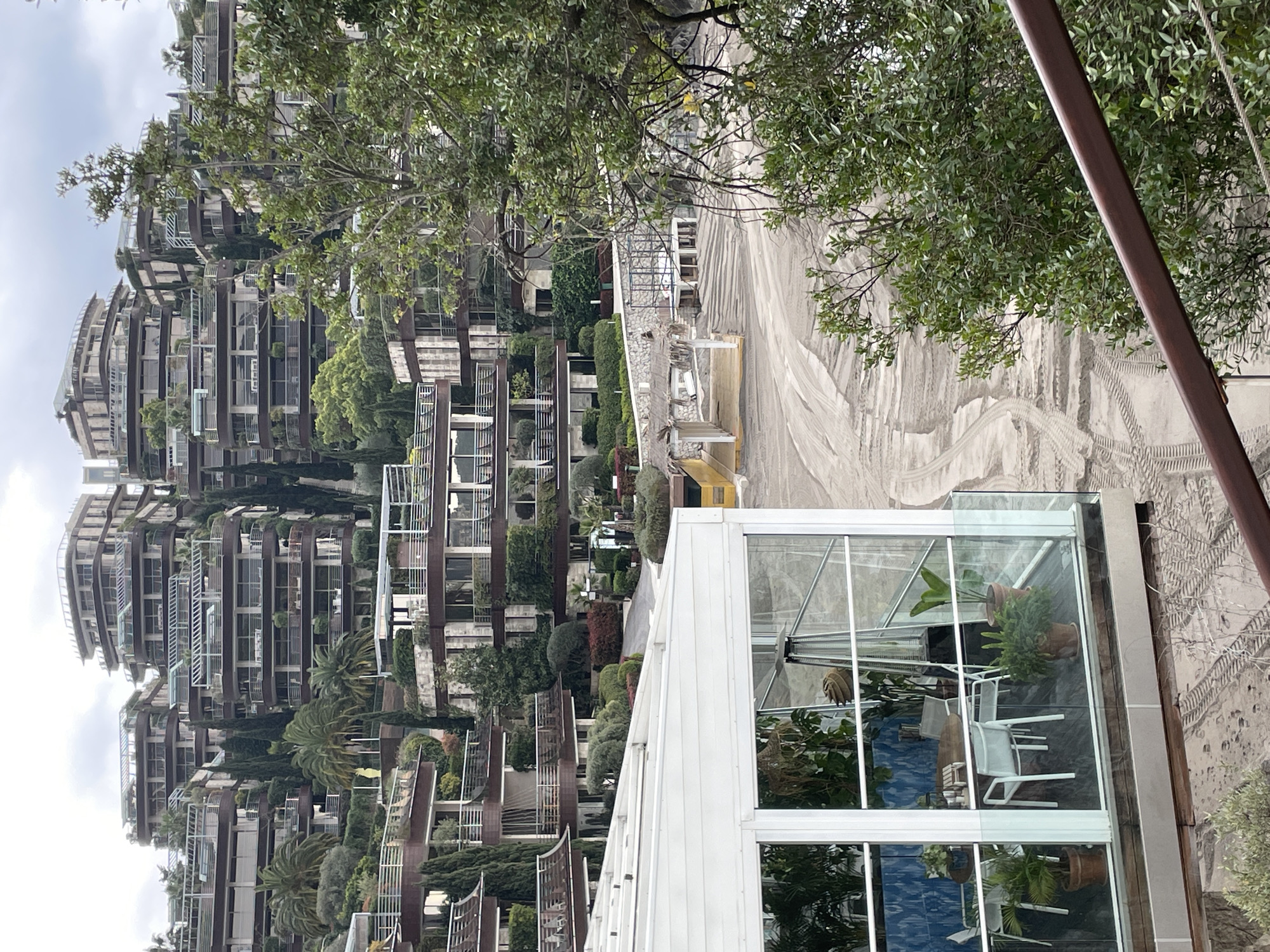 This image depicts a panoramic view of the Dukley Gardens residential and resort complex located on the Zavala Peninsula in Budva, Montenegro, under an overcast sky.
In the lower left foreground, a white, glass-enclosed structure, possibly a restaurant or cafe, stands on a dark deck. Visible through its large window panes are a round wooden table, four white chairs, several potted plants, and a freestanding silver outdoor heater. A glass balustrade extends from this structure, and a dark brown metal railing diagonally crosses the bottom right corner of the frame.
The midground features a wide, light-colored sandy beach exhibiting numerous tire tracks and disturbed patterns, indicating recent vehicle activity or landscaping. No individuals are present on the beach. On the left side of the beach, a small, open structure with a thatched roof and a wooden deck is visible. To its right, a white, skeletal framework on stilts, possibly a base for a temporary beach structure or platform, stands on the sand.
The background is dominated by a sprawling complex of terraced, multi-story buildings ascending a lush, green hillside. These modern structures feature dark brown facades, extensive curved balconies with white railings, and integrated garden elements on each level. The hillside is densely vegetated with numerous cypress trees, palm trees, and various green shrubs, interspersing and partially obscuring the buildings. The overall environment is one of an upscale coastal resort or residential area. No specific human actions or interactions are depicted.