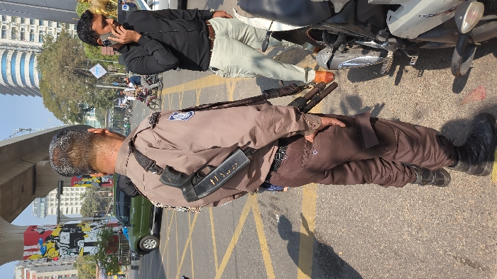 Two men stand on an urban street. One, in uniform, carries a rifle slung over his shoulder. He wears a brown jacket, dark brown pants, and black boots. A black pistol or other device is attached to his side. The second man wears a black shirt, light colored pants, and tan shoes. He is talking on a cell phone. A motorcycle is parked nearby. The background includes a building, a bridge, and traffic.