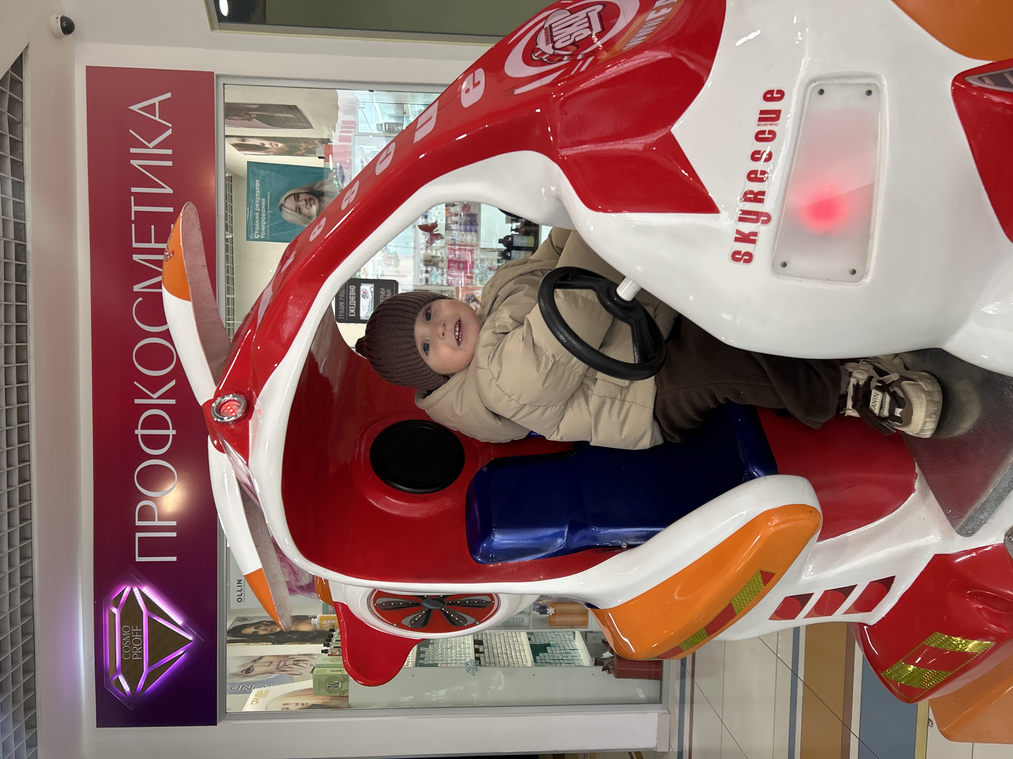 The image shows a child in a "skyrescue" airplane-shaped ride, likely located in a mall or shopping center. The child, wearing a brown hat and beige jacket, is positioned behind a steering wheel. The ride is painted in red, white, and orange, and features a propeller on the front. In the background, a store window displays "ПРОФКОСМЕТИКА" and the "COSMO PROFF" logo, with the text "OLLIN." There is also the statement "Стойкий результат Тонирования", and "ГРАФИК РАБОТ ЕЖЕДНЕВНО". Based on the Russian writing on store fronts in the background, the location appears to be in Klin, Russia.