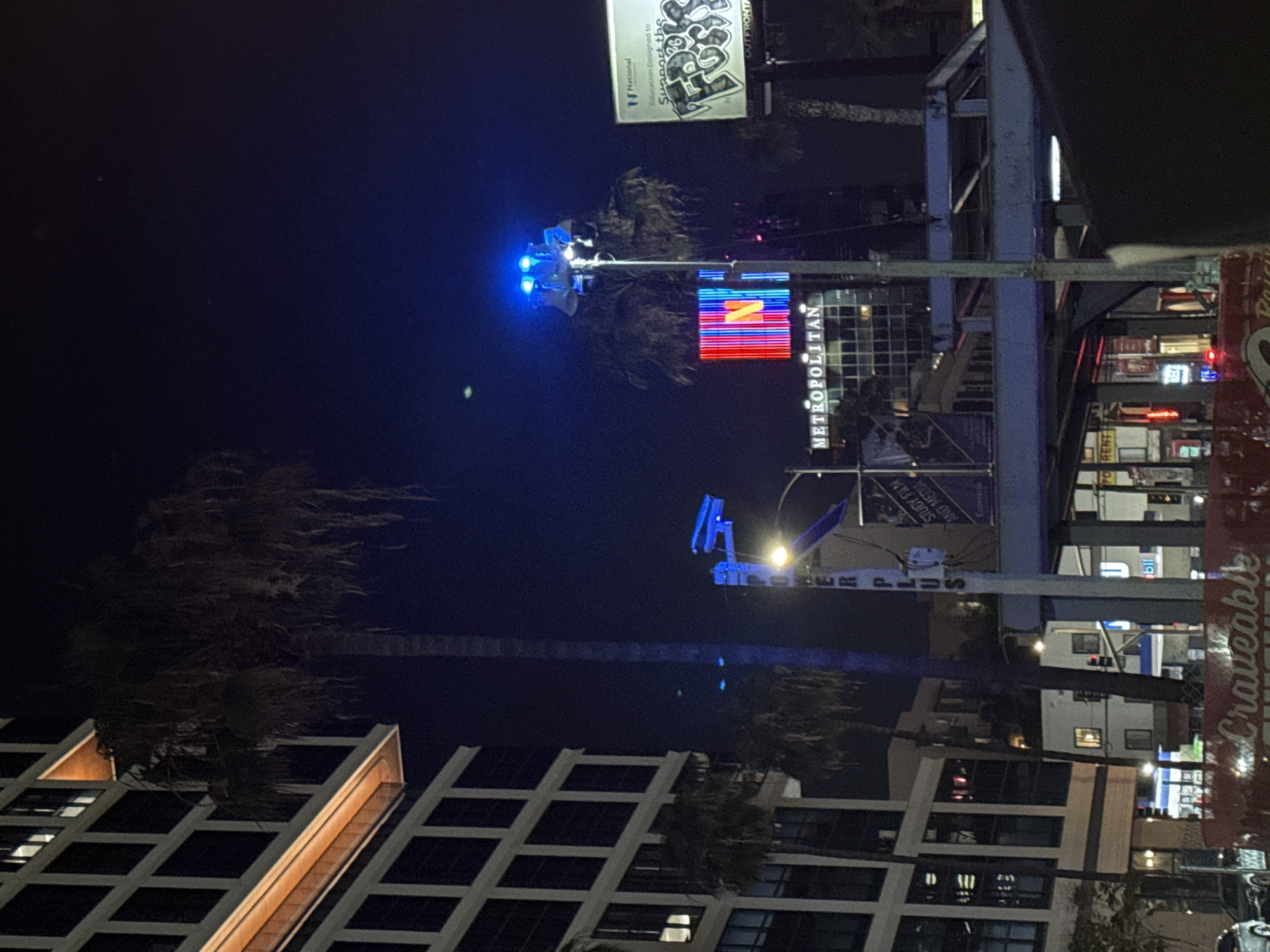 The image depicts an urban street scene at nighttime, characterized by a dark blue-black sky and a multitude of artificial lights illuminating buildings and signs. Several tall palm trees with dark, feathery fronds are visible, standing against the night sky and alongside the buildings.
In the mid-ground, a prominent street light or traffic signal assembly glows with an intense blue light. Below this, a large, colorful electronic sign displays abstract red, blue, and yellow patterns, with the word "METROPOLITAN" written vertically in white letters. Nearby, a billboard is partially visible, featuring text such as "National," "Education Designed to Support the," and "OUTFRONT," along with some graphic elements.
To the right, a structure with blue framing supports a banner or sign that reads "STUDY FILM AND MEDIA" and "Emerson." Below, the street level reveals various commercial signs and storefronts with internal lights, including partially visible text like "POWER PLUS," "bil," "Craveable," and "RENT." Modern multi-story buildings with grid-patterned facades and illuminated windows line the street.
The scene is devoid of people or discernible activity, presenting a static urban landscape. The time of day is clearly night, with clear weather. The presence of palm trees and the specific businesses and institutional names (like "Emerson") strongly suggest a city setting, consistent with Los Angeles, United States of America.