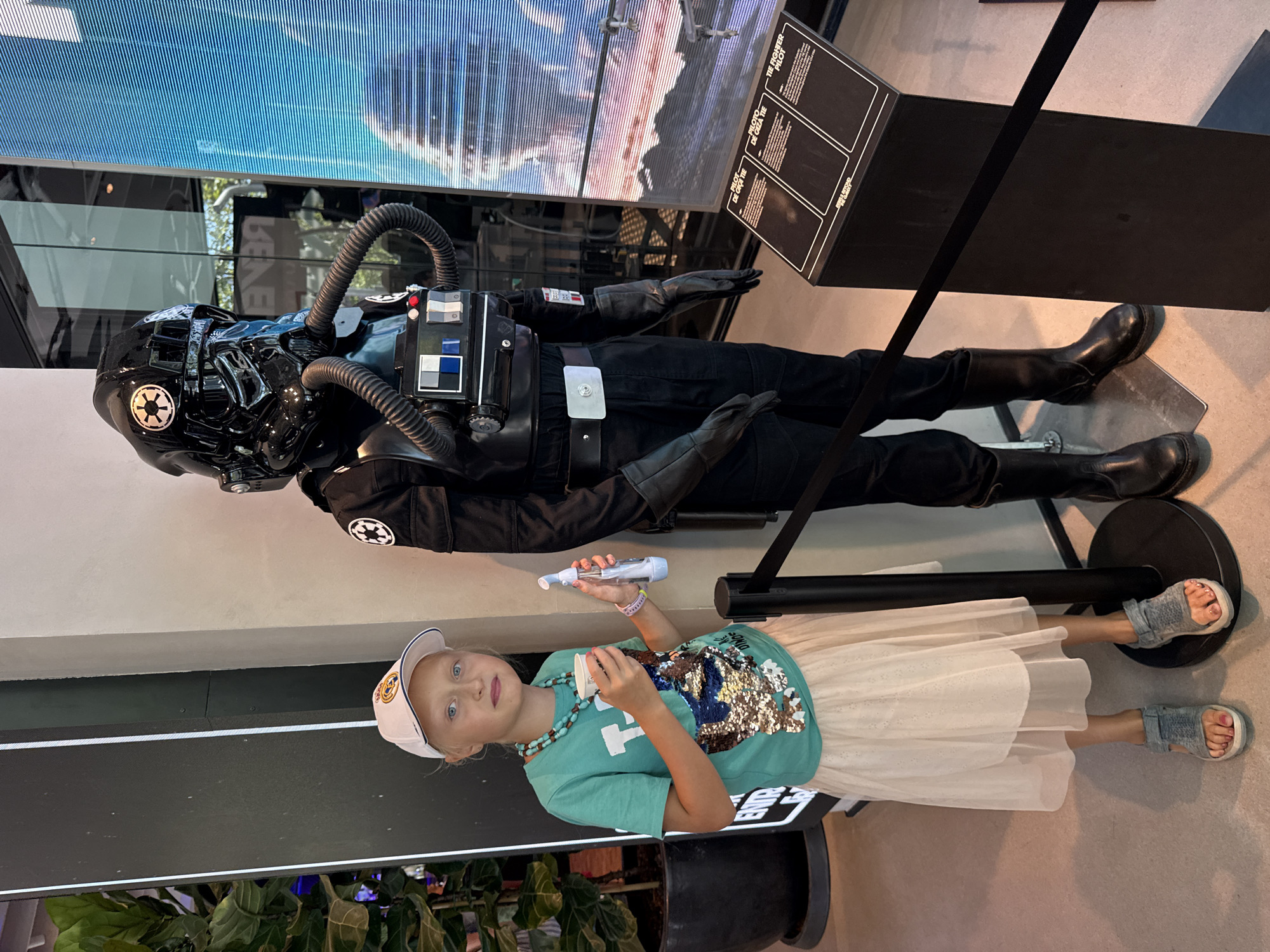 In Barcelona, Spain, a young girl stands to the left of a TIE Fighter Pilot mannequin from Star Wars. She is wearing a white baseball cap, a teal t-shirt with sequined embellishments, a sheer cream skirt, and grey sandals. She holds a small white cup in her right hand and a clear blue object in her left. The mannequin is dressed in a black TIE Fighter pilot uniform, complete with a helmet, gloves, and boots, and stands next to a tall, dark display stand with information in Spanish and English. Behind them, a large vertical screen displays a blue and white abstract pattern. The environment appears to be an indoor exhibit or attraction.