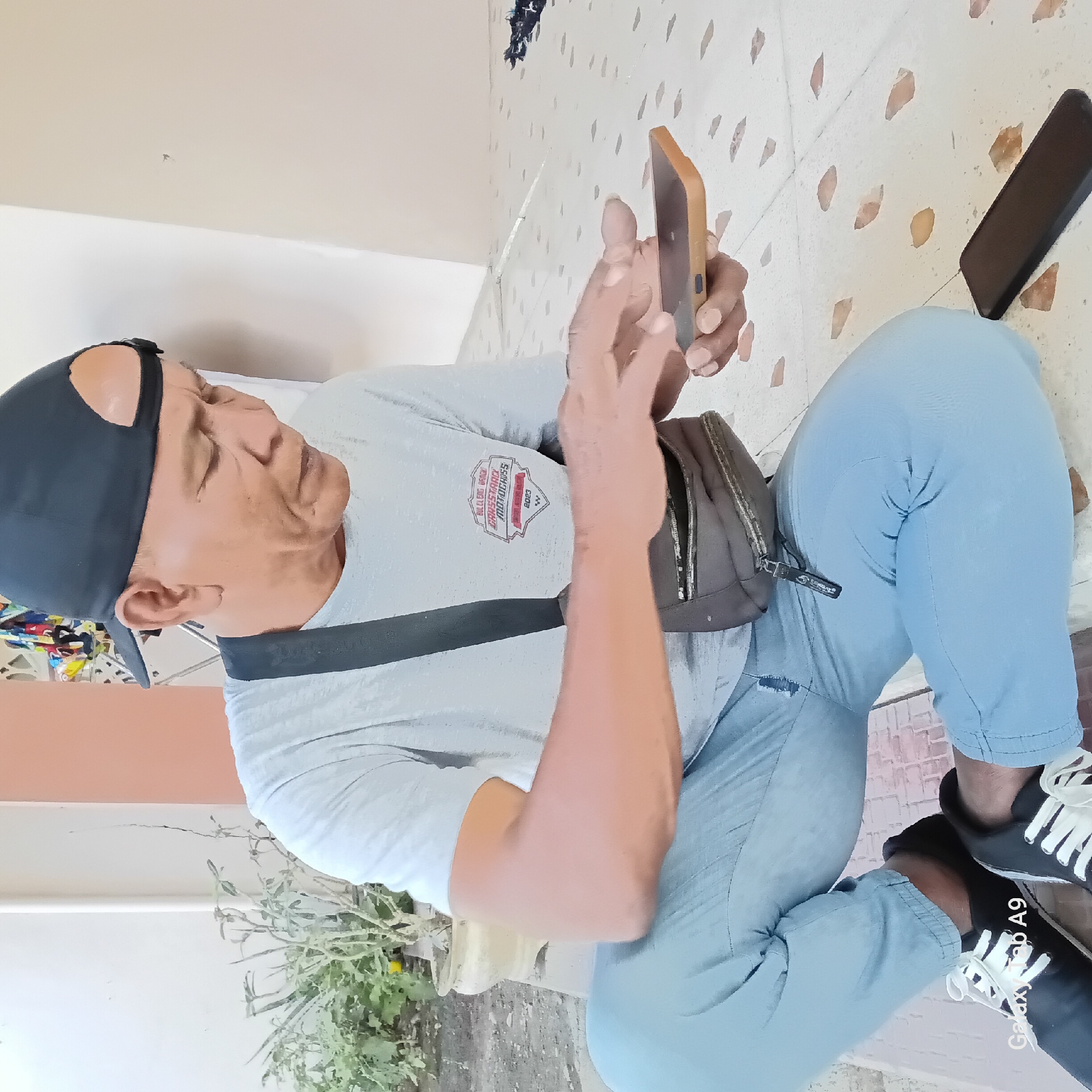 In Sambirejo, Indonesia, an older man wearing a grey t-shirt, light blue jeans, and a black baseball cap is squatting on a patterned tiled floor. He is looking down at his mobile phone, which he holds in his right hand, and appears to be touching the screen with his left index finger. A dark colored crossbody bag is slung over his shoulder, resting on his chest. Next to his right leg, a dark, rectangular object, possibly another phone or a power bank, lies on the floor. In the background, a potted plant with dry-looking foliage is visible, along with a light-colored wall and a section of a peach-colored wall. The overall setting suggests an outdoor or semi-outdoor area.