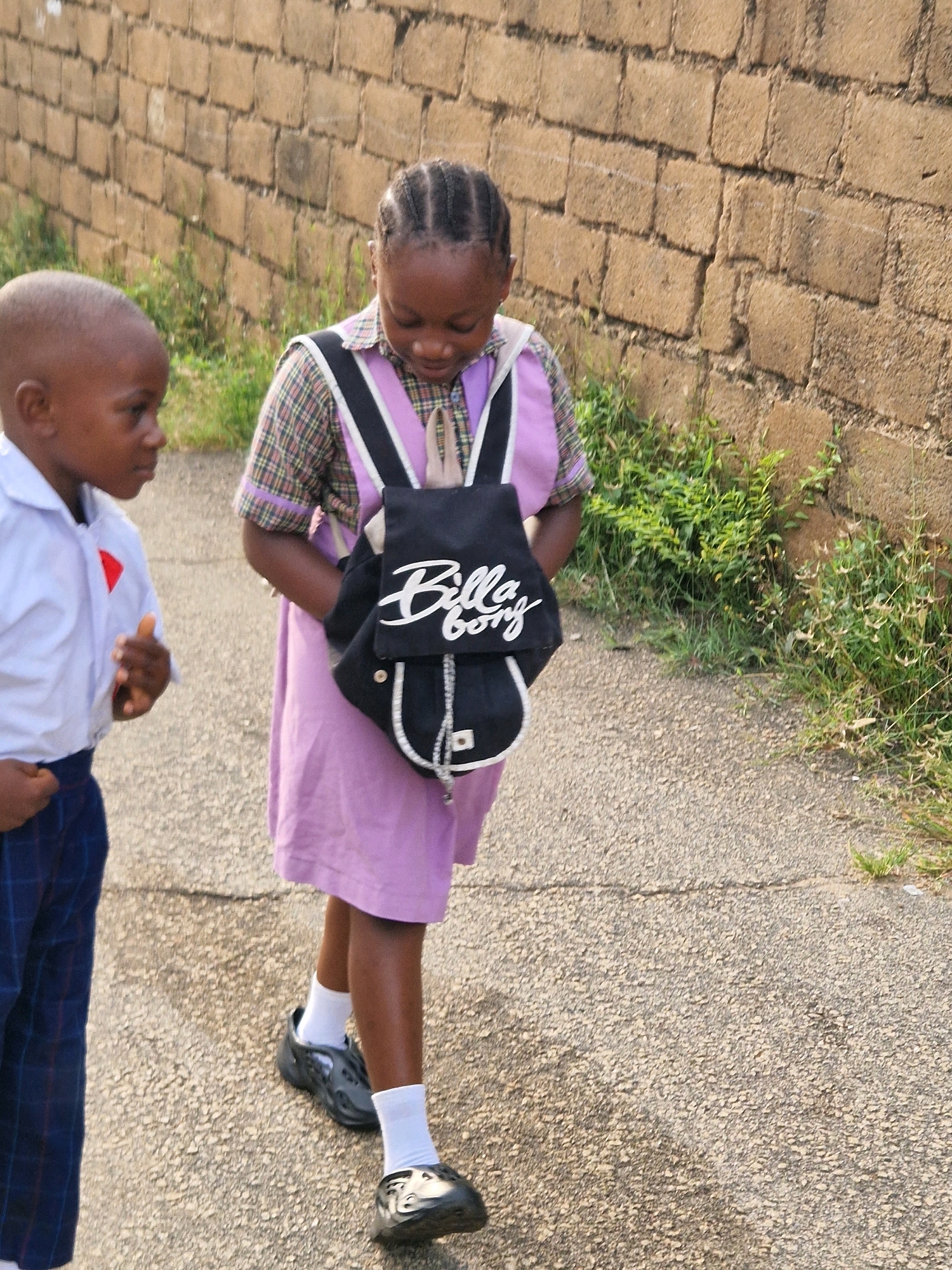 Two children are walking on a paved surface next to a brick wall in Kabong, Nigeria. On the left, a young boy wearing a white collared shirt with a red triangular emblem on the left side of the chest and dark blue checkered pants walks facing left. He has a shaved head. To his right, a girl wearing a purple dress and a plaid collar shirt walks facing forward. She has braided hair and is carrying a black backpack with white trim and "Billabong" written in white cursive on it. She is wearing dark shoes and white socks. The brick wall is made of rough, tan bricks and is behind the children. Green vegetation grows along the base of the wall. The paved surface is textured and grey, with some cracks visible. The lighting suggests it is daytime.