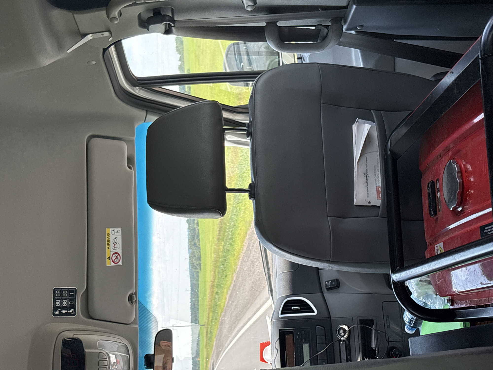The image depicts the interior of a vehicle, likely a van or bus, with a focus on the passenger seating area and dashboard. In the foreground, a red portable generator is visible, secured by black metal bars. To the left of the generator, a plastic water bottle sits on a green object, possibly a mat or bag.
Above the dashboard, a sun visor is partially visible, with an "AIRBAG" warning sticker and other symbols. On the ceiling of the vehicle, above the sun visor, a panel with multiple icons is affixed, indicating seating arrangements possibly related to passenger capacity or accessibility features, including icons for wheelchairs and seated individuals.
The driver's side of the dashboard is visible, showing the steering wheel and various controls. The view through the windshield reveals a rural landscape with a road, green fields, and trees under a cloudy sky. The interior of the vehicle appears to be clean and functional. The image does not contain any individuals. The image context suggests the vehicle may be in or traveling through the Мастоцкі сельскі Савет region of Belarus, based on the user prompt.