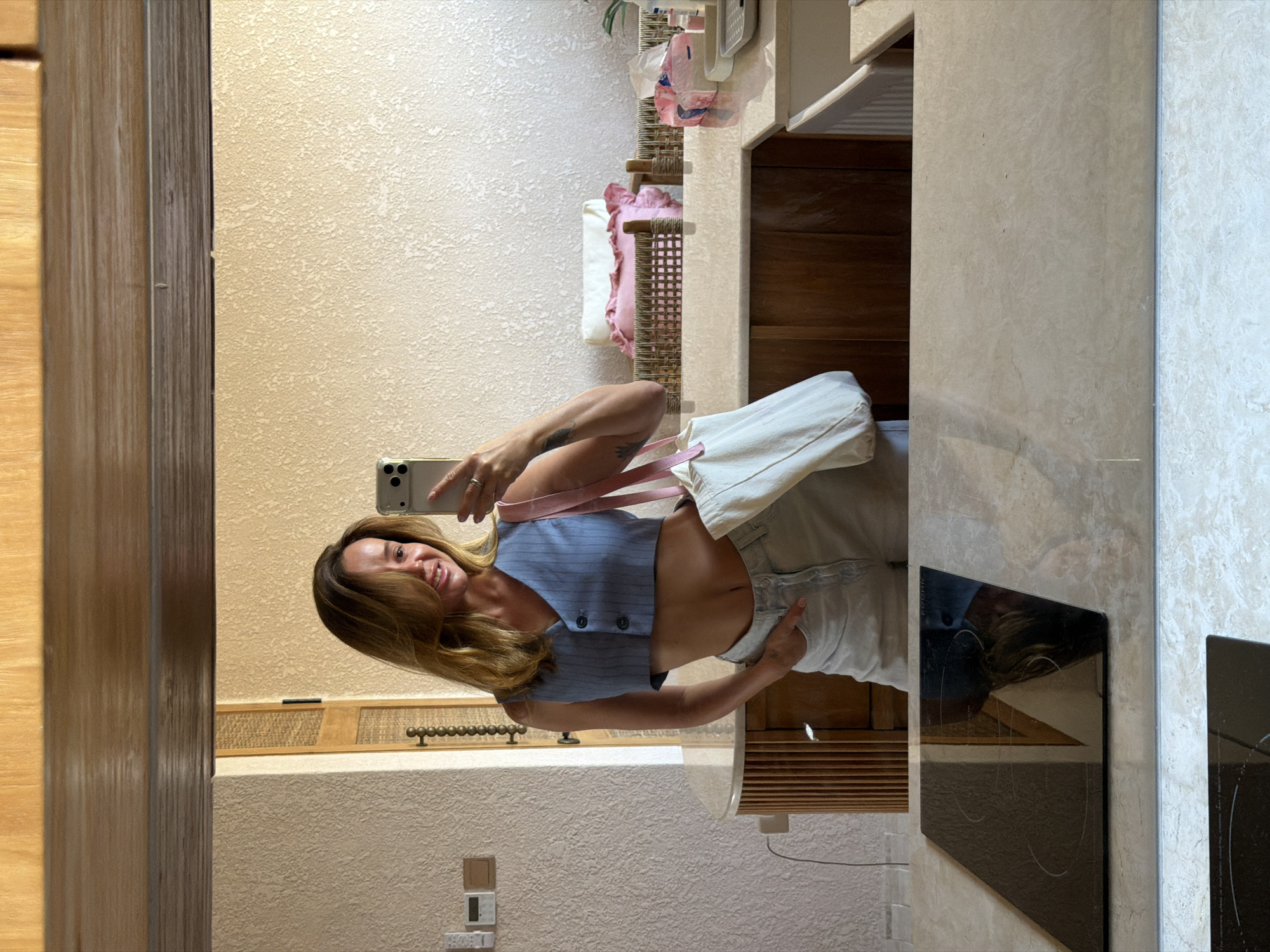 A woman is taking a mirror selfie in what appears to be a hotel room or a home. She is wearing a light blue, cropped vest over a white top and light-wash denim shorts. She is holding her phone in her right hand and a white tote bag with pink straps in her left. The background shows a textured wall, wooden cabinetry, a woven partition with pillows, and a countertop with various items. The scene is lit with natural light. The location in the city of Mengwi, Indonesia cannot be determined from the image.