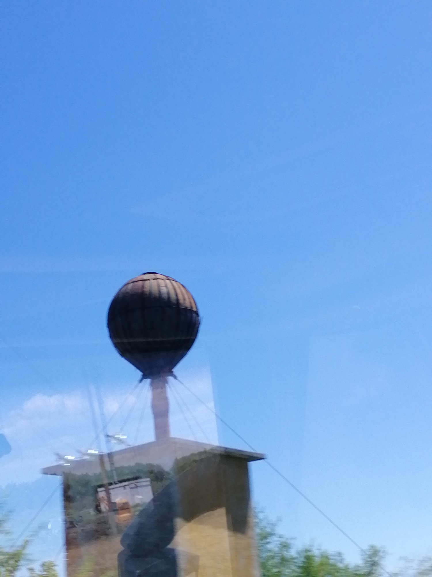 A water tower with a spherical, rusted metal tank stands against a clear blue sky. The tower is supported by a structure that appears to be made of concrete or stone, with some visible metal cables extending outwards. Below the water tower, a building or platform is visible, with greenery and trees surrounding it. There is a reflection in the foreground, suggesting the photo was taken through a window. The context of Durankulak, Bulgaria, is noted but not directly visible in the image itself.