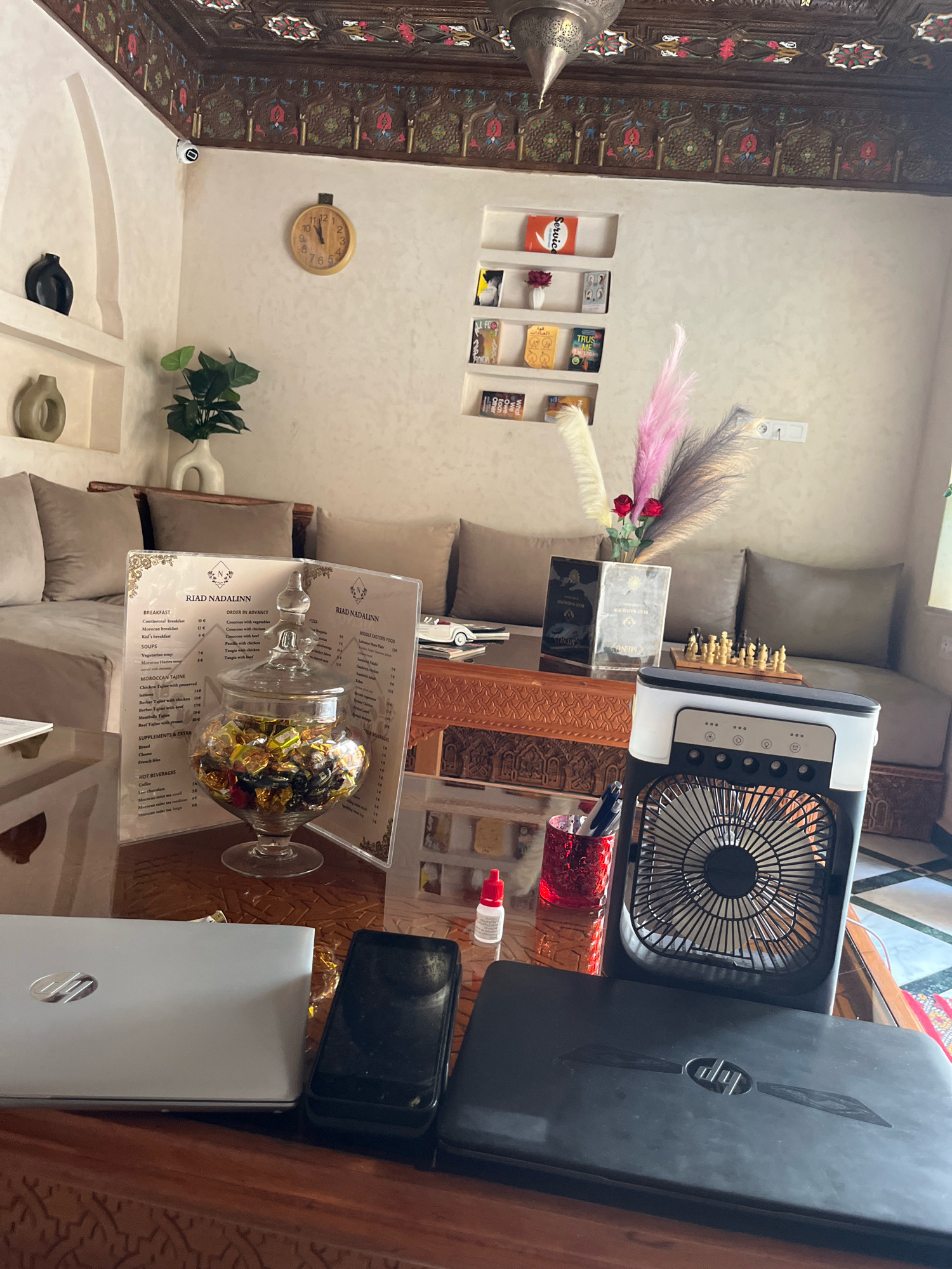 The image depicts an interior scene, likely within a riad or hotel in Marrakesh, Morocco, characterized by traditional Moroccan architectural elements and decor. In the foreground, a wooden table holds two laptops, one silver and one black, a smartphone, a small electric fan, a glass jar filled with wrapped candies, and a menu. A small red bottle and a red-patterned glass containing pens are also on the table.
Behind the table, a plush, upholstered sofa with multiple cushions dominates the mid-ground. The sofa is positioned in front of a intricately carved wooden screen that separates it from the rest of the room. On the wall behind the sofa, a decorative shelf displays several books and a small vase with flowers, accompanied by a round wall clock. Further to the left, recessed shelves feature decorative vases and a potted plant.
The ceiling is adorned with detailed wooden carvings and decorative patterns, typical of Moroccan craftsmanship. The overall ambiance suggests a comfortable and culturally rich setting. There are no people visible in the image, and the scene appears to be static, implying a captured moment in time.