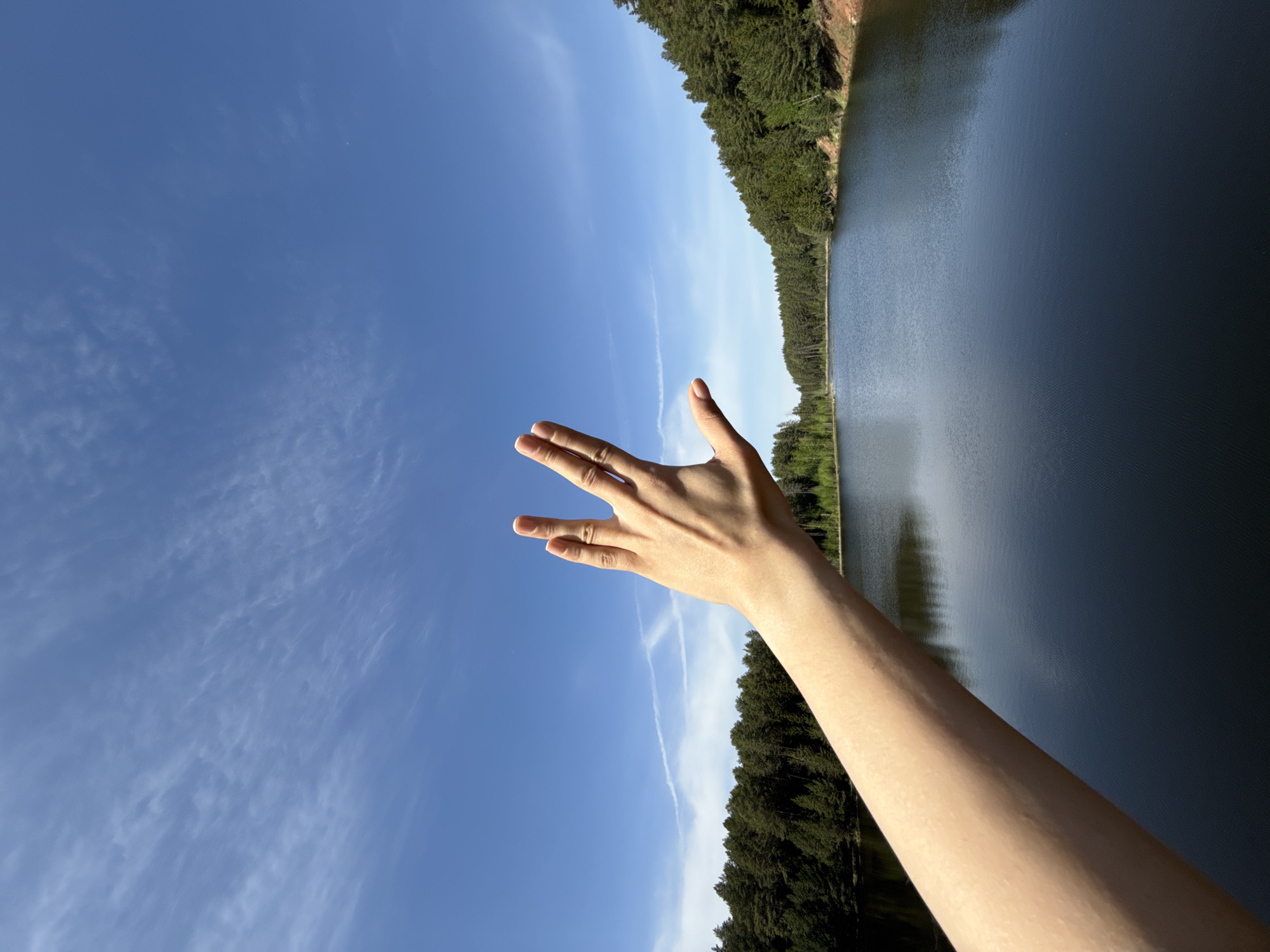 A human arm extends toward the sky in an outdoor setting. The hand is angled with the fingers spread apart. The background shows a clear, blue sky with wispy clouds. Adjacent to the sky is a body of water with a tree-lined shore. While the location is described as Votkinsk, Russia, there are no visual elements in the photo to confirm this.
