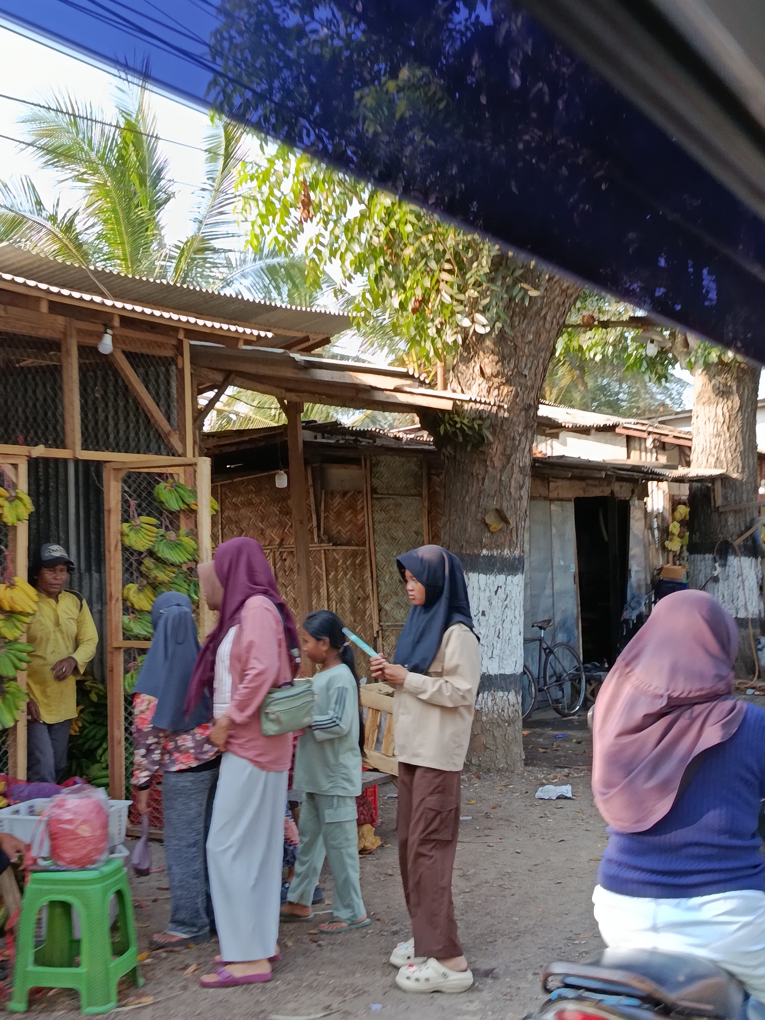 The image depicts a street scene in Ramban Wetan, Indonesia, with several individuals and market stalls. In the foreground, a person is seated on a motorcycle, viewed from the rear, wearing a dark blue top and white pants, with a maroon hijab. To the left, a young girl in a light green outfit with a white stripe is holding a light blue object. Beside her, a woman in a beige hijab and light brown pants stands, holding what appears to be a small item in her hands. Further left, another woman wears a pink hijab and a long white skirt, with a patterned fabric visible beneath her headscarf. Adjacent to her, a person in a dark blue hijab and a patterned top stands with a grey bag. To the far left, a man in a yellow shirt and a dark cap stands in front of a stall displaying bunches of green and yellow bananas. Various market structures made of wood and corrugated metal are visible in the background, with bamboo weaving incorporated into some walls. A bicycle is parked near a wooden stall in the mid-ground. The ground is dirt, and trees are present. The scene appears to be a marketplace, likely in the morning or late afternoon, given the lighting.
