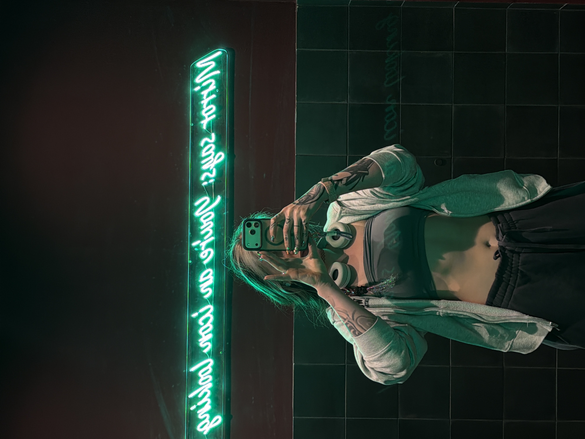 A woman with tattoos on her arms is holding a phone and taking a picture of herself in front of a green neon sign. She is wearing a sports bra, sweatpants, and a hoodie. She is also wearing headphones around her neck. The background is a dark red wall and a black tiled wall. There is writing in green neon light. The location cannot be determined from the image.