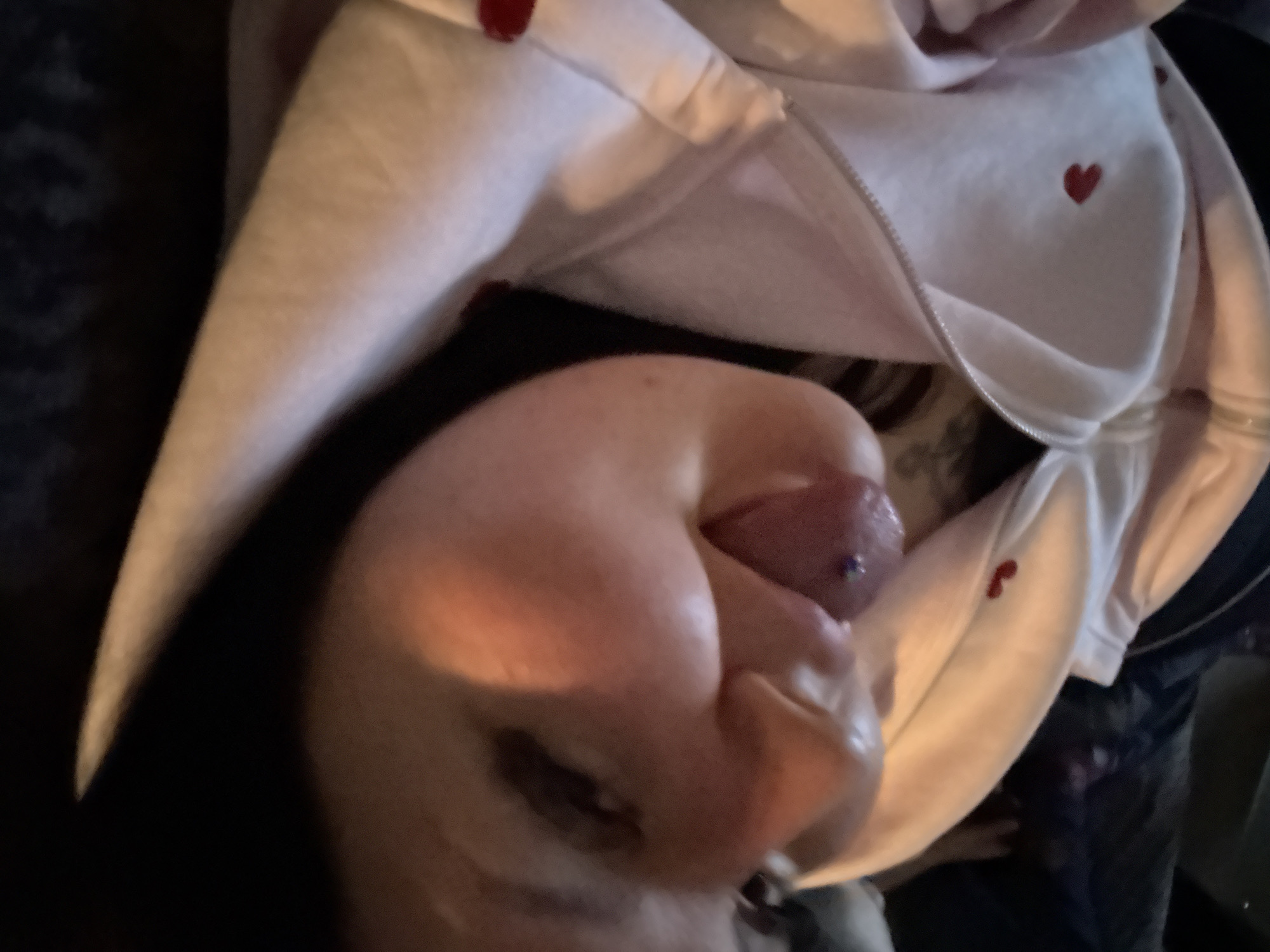 A person is visible, lying down and facing the camera. The person has a pierced tongue. They are wearing a light-colored hooded sweatshirt with red heart designs. The location is unknown and cannot be determined from the image.