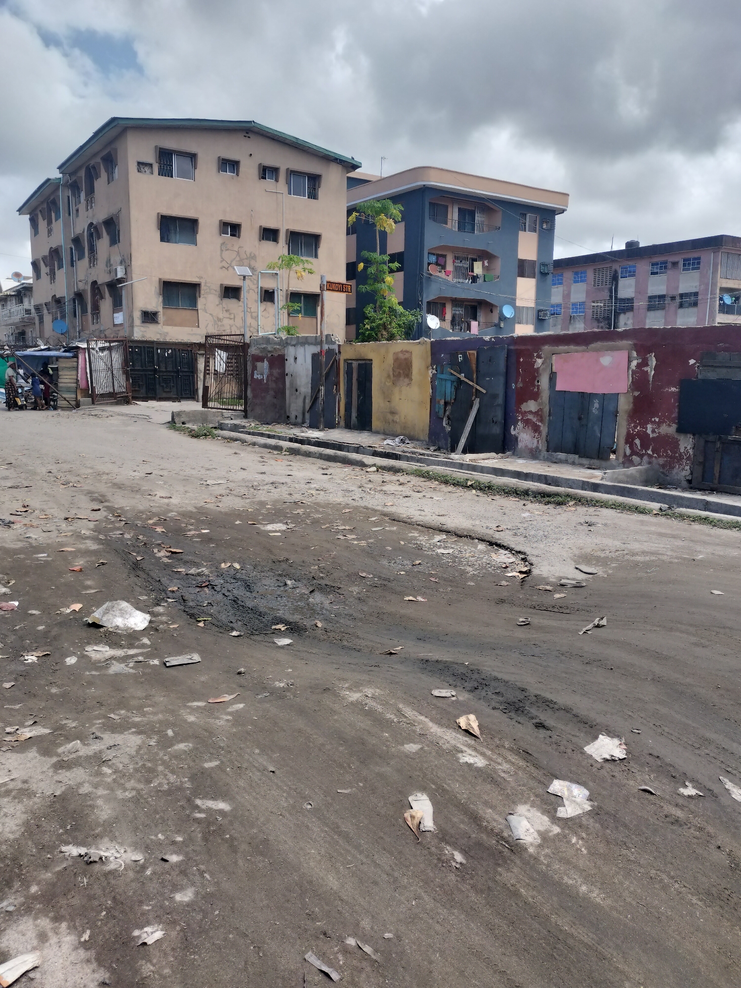 The image depicts a street in Alaiyabiagba, Nigeria. The road surface is degraded and covered in debris. Along the roadside, there are buildings of 2-4 floors and shacks or stalls constructed. The sky is overcast. There are a few people in the distance near a group of motorbikes. A street sign reading "KUKOYI STR" is visible.