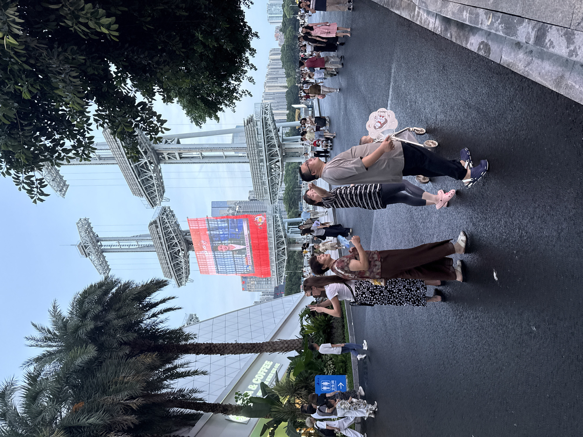 In this vertical, wide-angle shot, a crowd of people is gathered on a paved outdoor promenade. The time appears to be late afternoon or early evening, with overcast skies and muted lighting. The scene is set in a modern urban environment, with tall buildings visible in the background and a large metallic structure, possibly part of a stadium or event venue, dominating the mid-ground. Lush green trees, including palm trees, line the left side of the frame, adding a touch of nature to the cityscape.
Several individuals are the primary focus of the image. In the foreground, a man wearing a grey t-shirt and dark pants holds a white fan decorated with a cartoon cat. He is accompanied by a woman in a striped dress and leggings. Further down the promenade, a woman in a patterned top and dark pants is looking at her phone, while another woman in a white t-shirt and a patterned skirt is also engaged with her device. More people are visible further down, forming a queue or gathering, suggesting an event or popular attraction. The overall mood seems relaxed and casual.
There is visible text on a red banner hanging from the metallic structure, displaying an image of a man and Chinese characters. Additionally, a sign for "COFFEE" is visible on a building in the lower part of the frame. The setting in Tianhe District, China, is hinted at by the urban architecture and the presence of a specific type of palm tree often found in warmer climates.