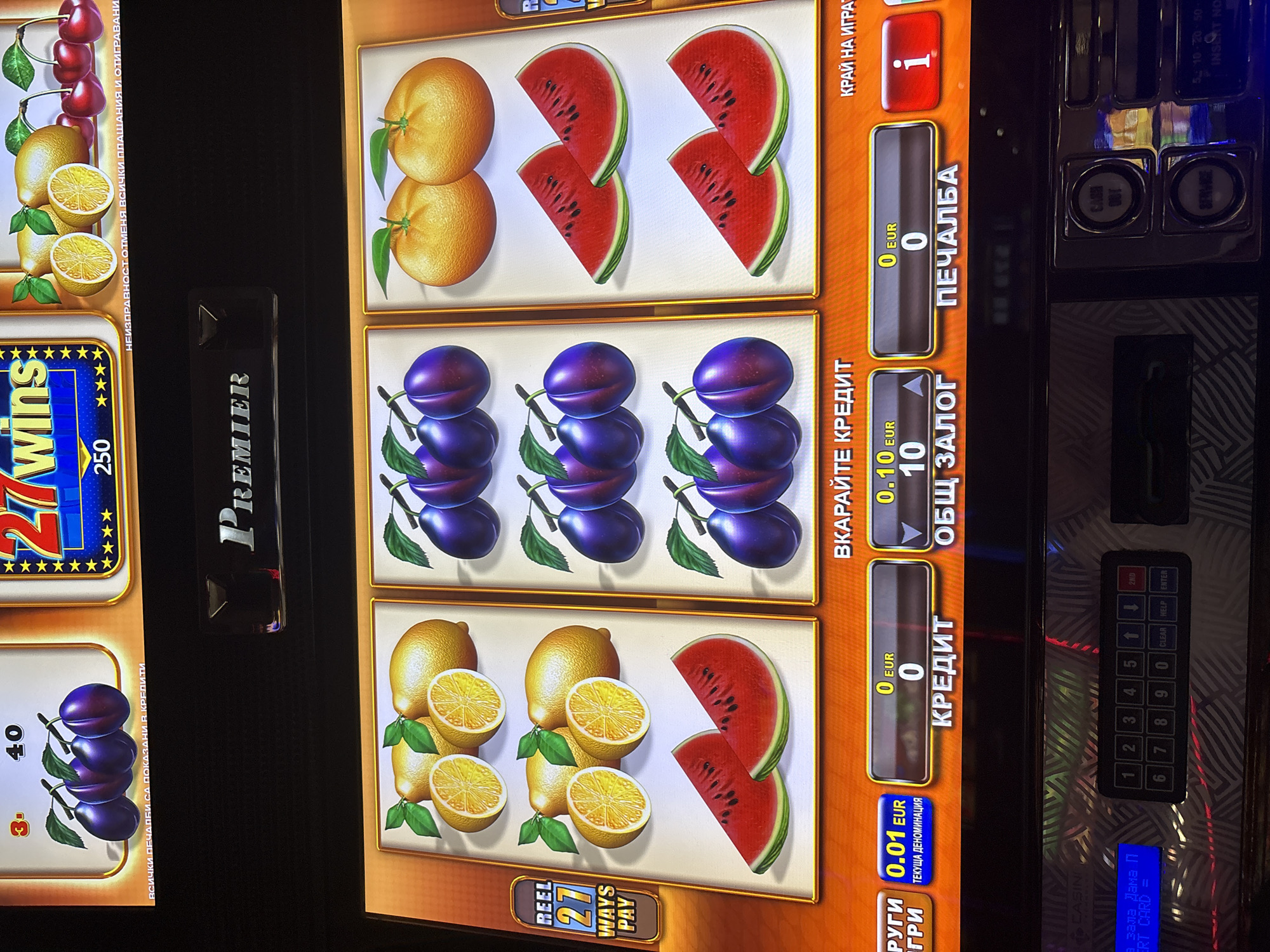 The image shows a close-up of a slot machine with colorful fruit symbols like plums, lemons, cherries, and watermelons. The machine displays various information in what appears to be Bulgarian text, including "РЕЕЛ 27 WAYS PAY", "ВКАРАЙТЕ КРЕДИТ", "КРЕДИТ", "ОБЩ ЗАЛОГ", and "ПЕЧАЛБА". There are also numerical values and currency indicators like "0.01 EUR" and "10 EUR". A keypad and some buttons are visible at the bottom of the machine. The overall environment appears to be an indoor gaming or entertainment venue. The image itself does not contain any direct information about the city of Karlovo, Bulgaria, nor are there any people or specific actions depicted beyond the context of the slot machine's operation.