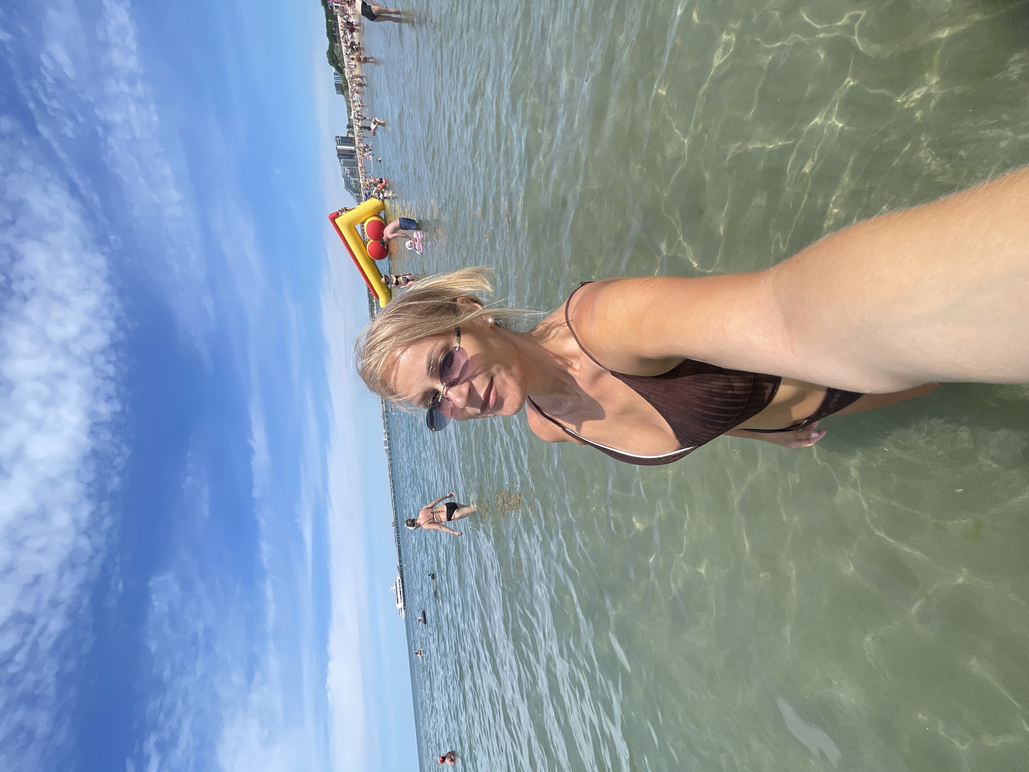 A woman is taking a selfie in the ocean. She is wearing a brown bikini and sunglasses, with her blonde hair pulled back. The water is clear and shallow, with sunlight glinting off the sandy bottom. In the background, a crowded beach stretches out with people sunbathing and swimming. An inflatable water slide with red and yellow sections is visible in the water near the shore. Buildings and trees line the coastline in the distance. The sky is bright blue with scattered white clouds.