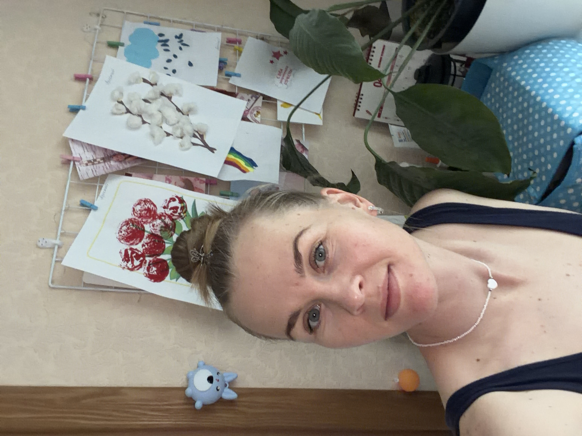 This is a medium shot, eye-level, indoor selfie of a woman in her late 20s or early 30s. She has fair skin, blue eyes, and her brown hair is pulled up in a bun. She is wearing a dark blue sleeveless top and a white beaded necklace with a heart-shaped pendant. She is standing in front of a wall decorated with several children's drawings clipped to a wire grid. The drawings depict a bouquet of red flowers, branches with white cotton-like tufts, a blue cloud with raindrops, and a rainbow. To the left of the woman, a small blue cartoon character toy is attached to the wall. To the right, a large green plant with broad leaves is visible, and behind it, a desk calendar is partially obscured. A light wood door frame is visible on the left side of the image. The scene suggests a home environment, potentially a child's room or a creative space within a home. The image does not provide specific visual cues that can place it in Khabarovsk, Russia.