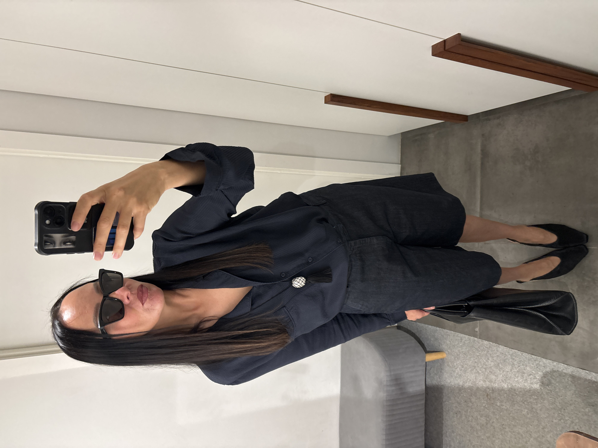 A woman with long, dark hair and sunglasses is taking a selfie in what appears to be a dressing room or fitting room. She is wearing a dark-colored, button-down shirt with a decorative button and tassel detail, along with what seems to be a matching pair of shorts or culottes. She is holding a black smartphone in her left hand, and in her right hand, she holds a black leather tote bag. The woman is also wearing black shoes, possibly ballet flats. She is sitting on a light gray bench.
The background includes a white wall with a horizontal light gray line and a white wall with two wood molding accents. The floor is concrete.
There is no information to conclude that this is in Chaikovsky, Russia.