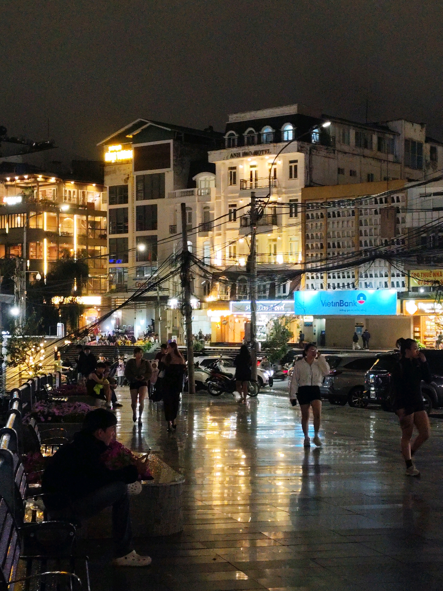 The image captures a rainy evening scene on a street in Da Lat, Vietnam. The street is wet, reflecting the brightly lit buildings and streetlights, creating a vibrant and somewhat surreal atmosphere.
In the foreground, a man sits alone on a bench, facing away from the camera, holding what appears to be a bouquet of flowers or foliage. He is wearing a dark hoodie and light-colored Crocs. To the right of the frame, several women are walking on the wet pavement. One woman, in the mid-ground, wears a white jacket and dark shorts, looking ahead. Another woman in the right mid-ground wears dark clothing and shorts, her back to the camera. Further back, other pedestrians are visible, some walking and others gathered in a small group.
The background is dominated by multi-story buildings, characteristic of an urban environment. Some buildings are illuminated with warm yellow lights, suggesting shops or residential spaces, while others are a lighter color. A prominent blue sign displaying "VietinBank" is visible, indicating the presence of a bank. The sky is dark and cloudy, typical of an evening with rain. Overhead, a complex network of electrical wires is visible, a common sight in many Vietnamese cities. The overall impression is a bustling city street transitioning into nighttime, with the rain adding a reflective quality to the environment. The specific location is identifiable as the vicinity of Anh Dao Hotel on Trieu Viet Vuong street in Da Lat, a known tourist area.