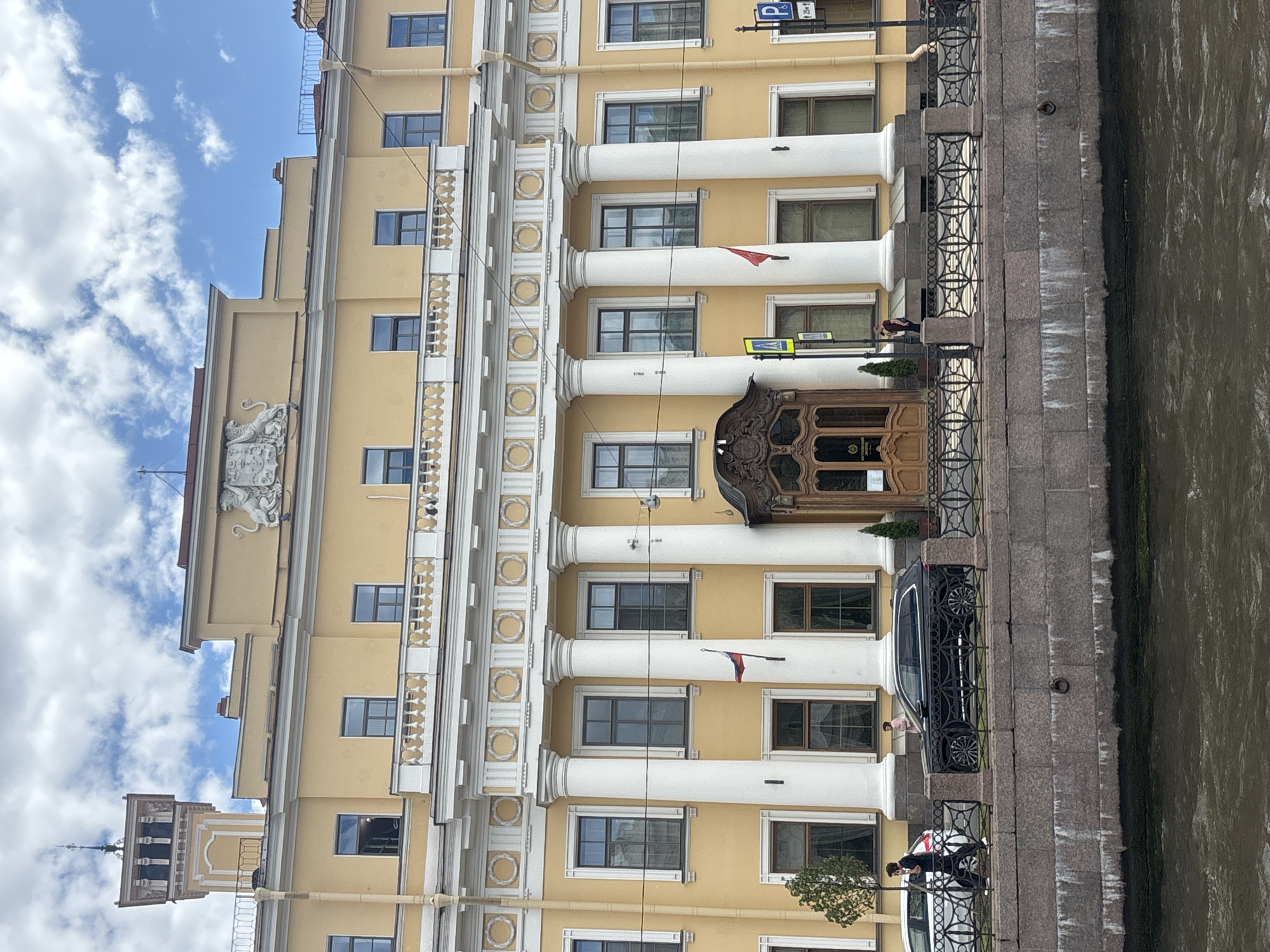 The image shows a grand yellow building with white columns and ornate architectural details, located in Saint Petersburg, Russia. The building faces a canal, with a stone embankment and an decorative metal fence separating the building from the water. The building's facade features multiple windows, a large wooden entrance with intricate carvings, and a Russian flag flying from a flagpole. Above the entrance, a coat of arms is visible. On the street level, a few people are walking, and a black SUV and a white car are parked. The sky is partly cloudy.