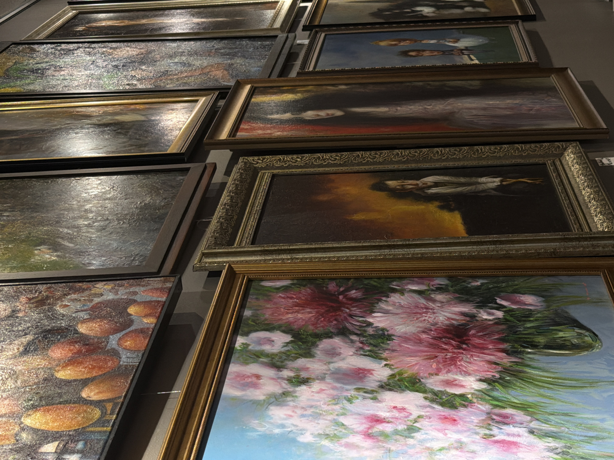 The image shows a collection of paintings displayed on a wall, likely in an art gallery or exhibition space. The paintings vary in subject matter and style.
In the foreground, a large, ornate golden-framed painting depicts a vibrant still life of pink and red flowers in a glass vase against a light blue background. To its right, another painting, framed in ornate dark wood, shows a portrait of a woman with dark hair and a pale face, rendered in a somber, almost ghostly style. To the right of that, a painting with a golden frame features two children, one in a white dress and the other in a blue dress, against a dark background. Further to the right, a painting with a darker frame shows a black and white cat.
Above the floral still life, a landscape painting with a textured, impasto technique is visible, featuring warm tones of orange and yellow, possibly depicting fruit or abstract shapes. To its right, a large, rectangular painting with a dark frame displays a dark, moody landscape with muted colors. Other paintings, partially visible, suggest a diverse range of artistic expressions.
This scene is located in the State Hermitage Museum in Saint Petersburg, Russia. The museum, one of the largest and oldest museums in the world, houses an extensive collection of art and cultural artifacts, including a vast array of paintings.