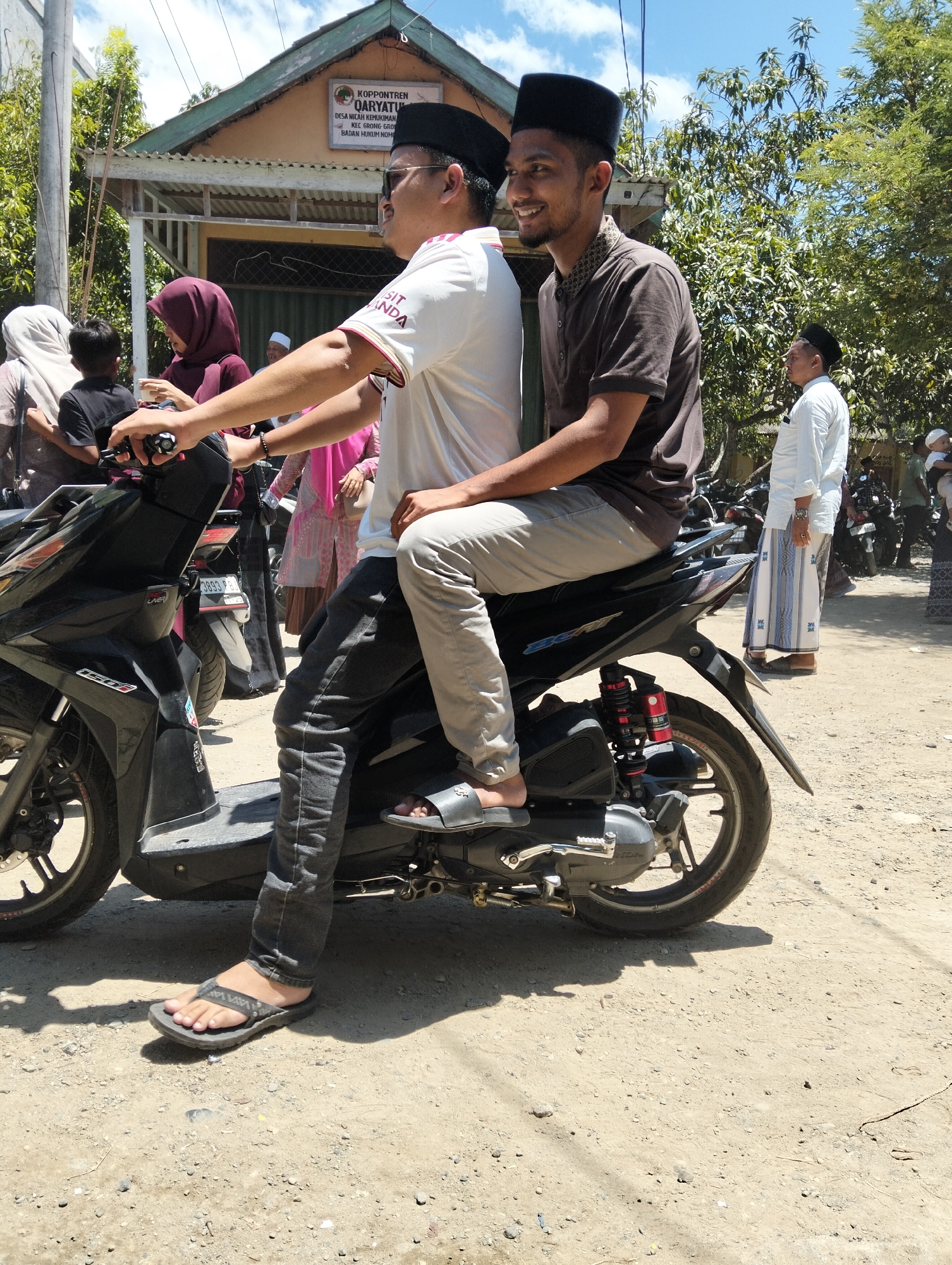 Two men are riding on a black scooter in the city of Teungoh, Indonesia. The man in the front is wearing a white t-shirt with red trim and black pants, and the man behind him is wearing a brown t-shirt and light gray pants. Both men are wearing black skullcaps. They are on a dirt road in front of a building with a sign that reads "Koppontren Qaryatun." There are other people and motorcycles in the background, and trees and a blue sky with clouds overhead. The lighting suggests it is daytime.