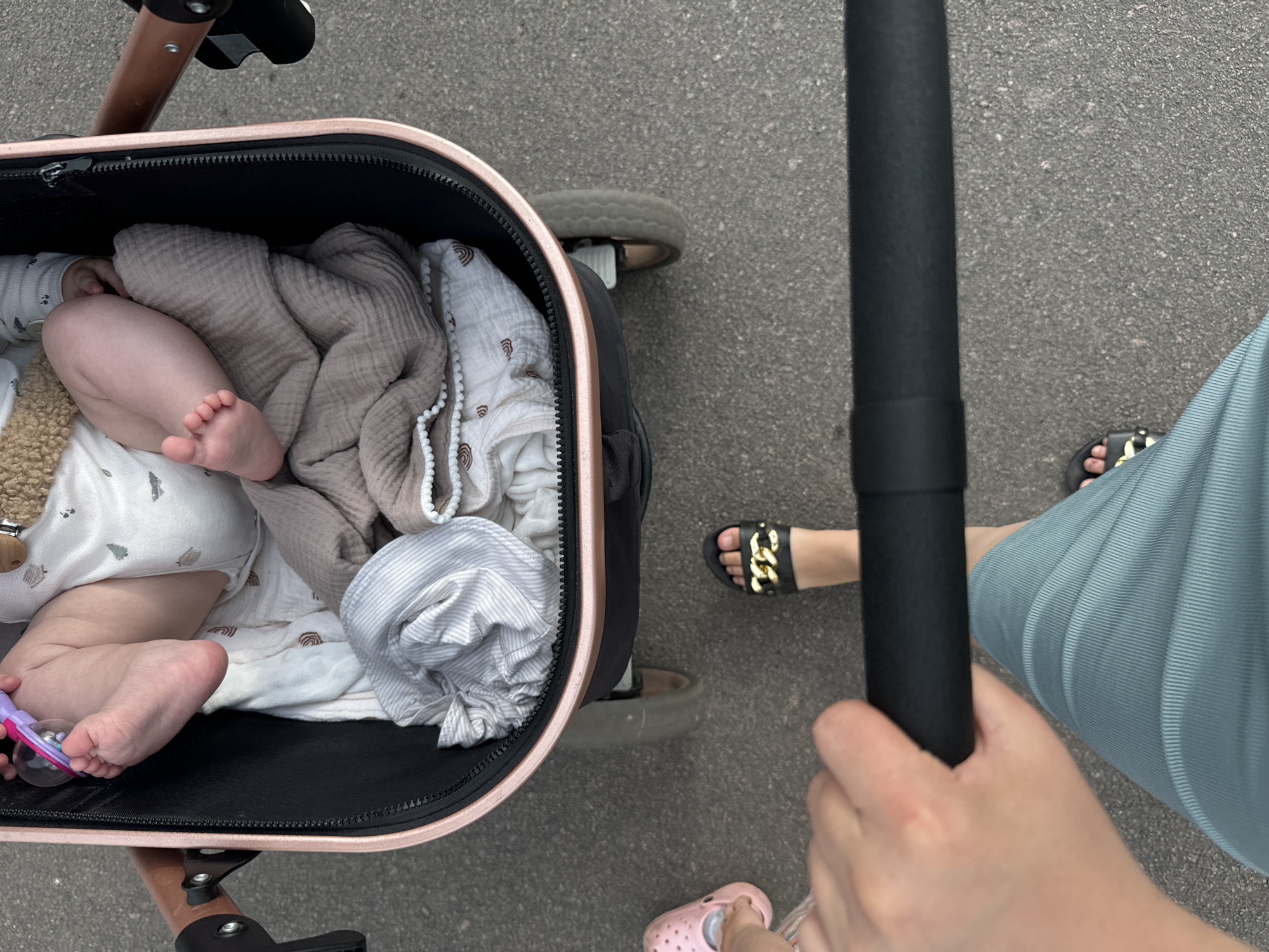 The image shows a first-person perspective of someone pushing a stroller. In the stroller, a baby's legs are visible, along with blankets and a pacifier. The stroller is a dark color with rose gold accents. The ground appears to be asphalt. A person's hand is visible gripping the stroller handle. Another person's feet, wearing pink slides and black sandals, are also in the frame. The scene suggests a walk in an outdoor urban environment. The location is specified as Dovhe, Ukraine.