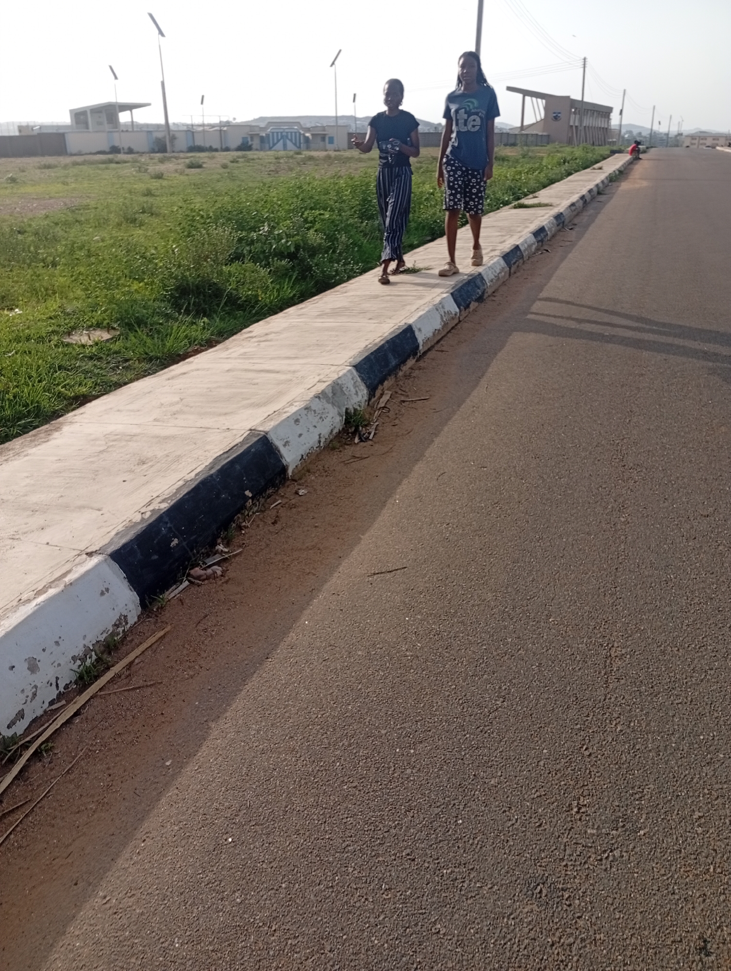 Two young women are walking on a paved sidewalk along a street in Jos, Nigeria. The woman on the left is wearing a dark blue t-shirt and striped pants, while the woman on the right is wearing a dark blue t-shirt with a graphic design and patterned shorts. They are walking side-by-side, with the woman on the left looking towards the right. Behind them, there is a grassy area with some buildings and streetlights in the distance. The street is asphalt and appears to be a relatively new development. The weather is clear and sunny.
