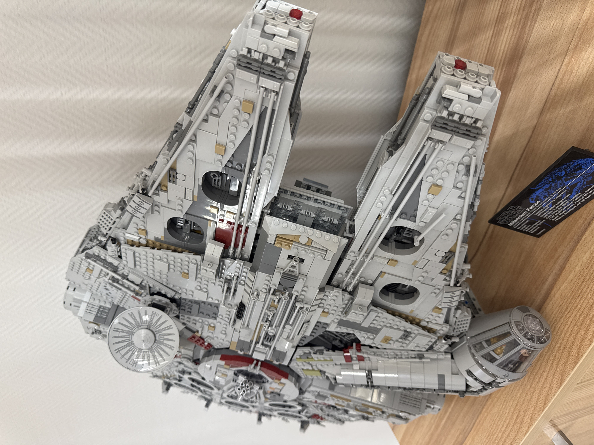 This is a close-up, angled shot of a Lego Millennium Falcon model. The spacecraft, constructed from gray and white Lego bricks with subtle tan and red accents, dominates the frame. Details like the radar dish, cockpit, and intricate surface detailing are visible. The model rests on a surface with a wooden edge visible on the right. To the right of the model, a small black card featuring the "Star Wars" logo and some text details the Millennium Falcon. The background is a plain, light-colored surface. There's no indication this image was taken in Krasnoyarsk, Russia. The available information solely describes the Lego model and its immediate surroundings.